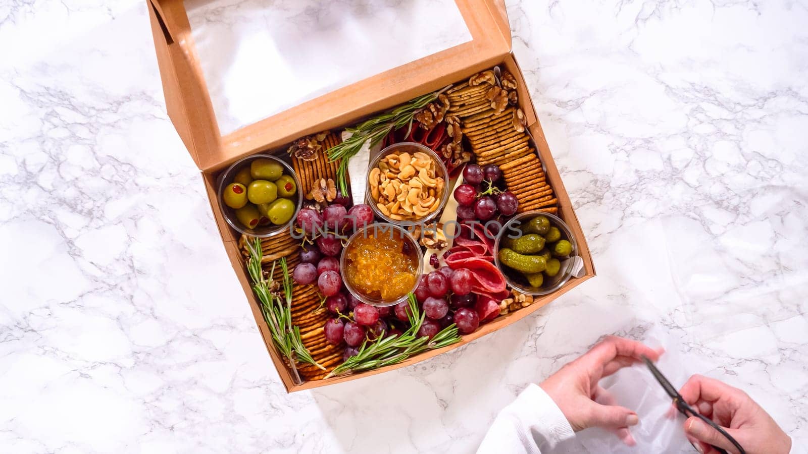 Flat lay. Hands are meticulously adding fresh red grapes to a bowl, complementing a beautifully arranged charcuterie box brimming with a variety of cheeses, olives, and cured meats, set against a sleek marble surface.