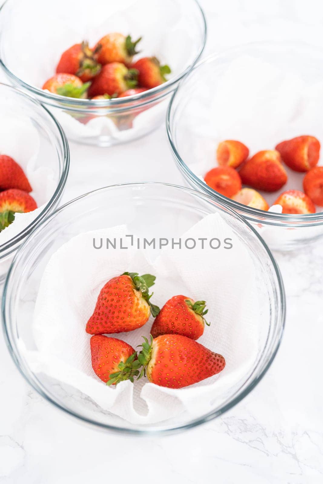 Bright red strawberries, interspersed with signs of mold, rest in a glass bowl lined with a paper towel on a white napkin, indicating improper storage techniques.
