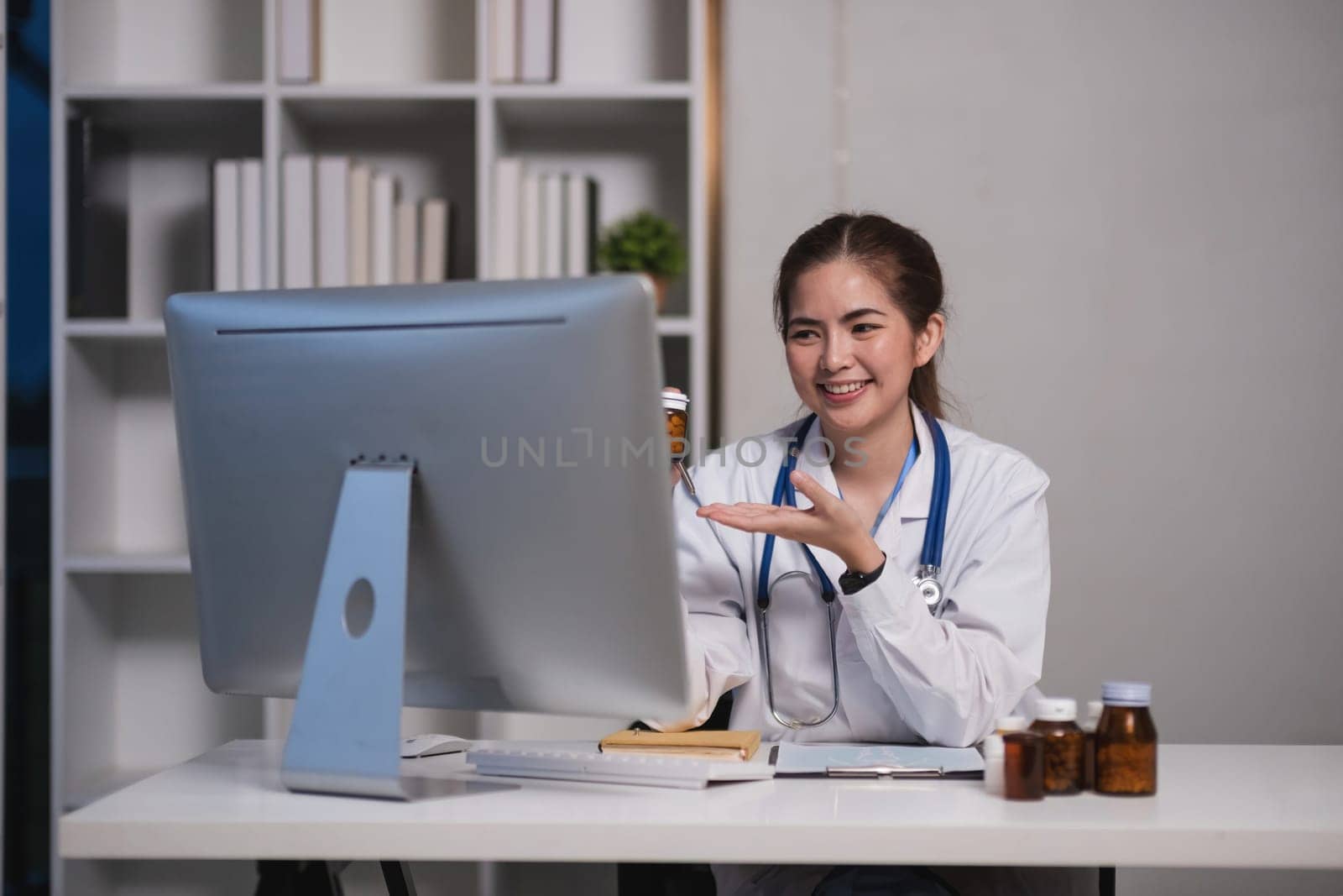 A woman in a white lab coat is sitting at a desk in front of a computer monitor. She is smiling and she is happy