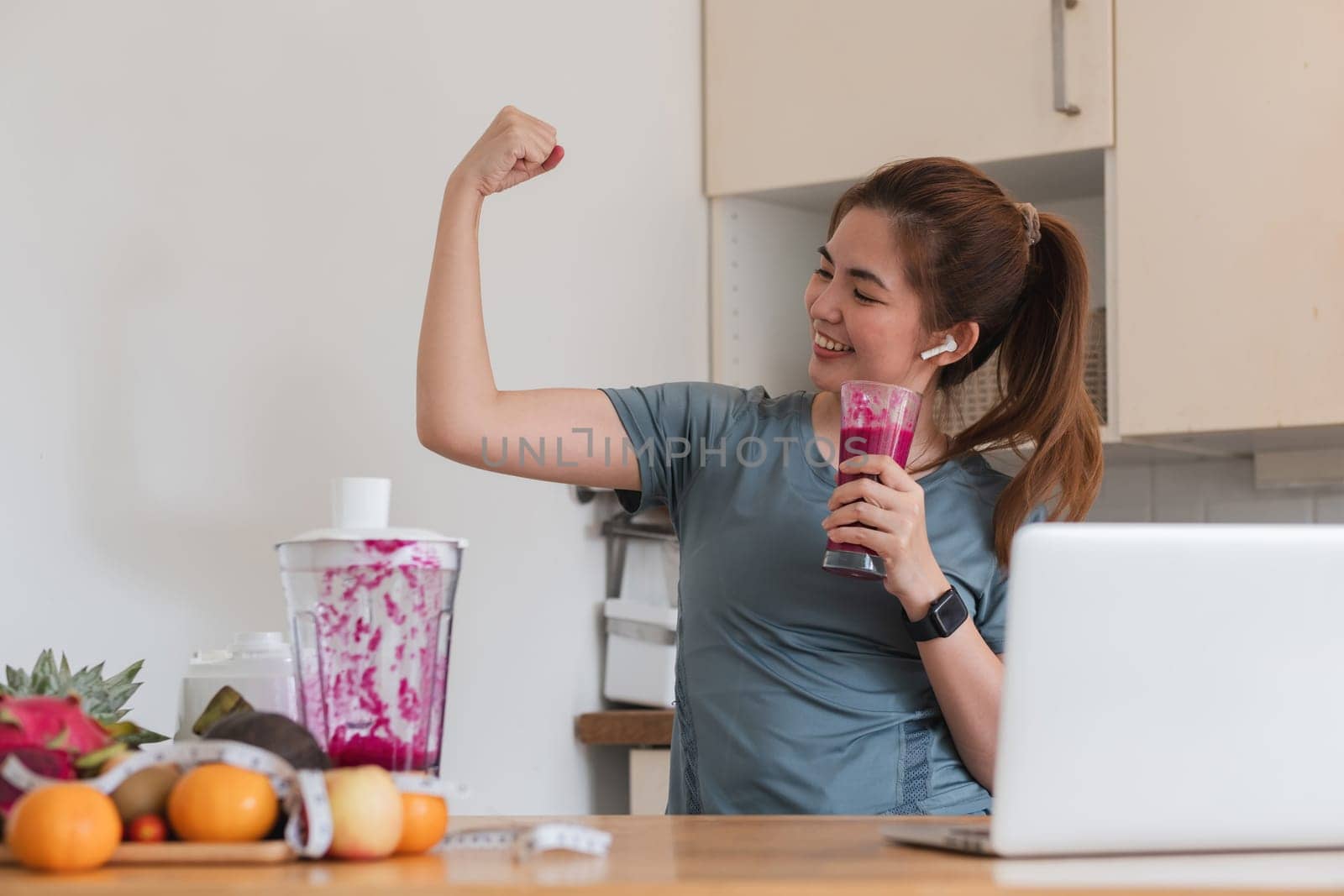 A woman is making a smoothie and is smiling. She is wearing a blue shirt and has a watch on her wrist