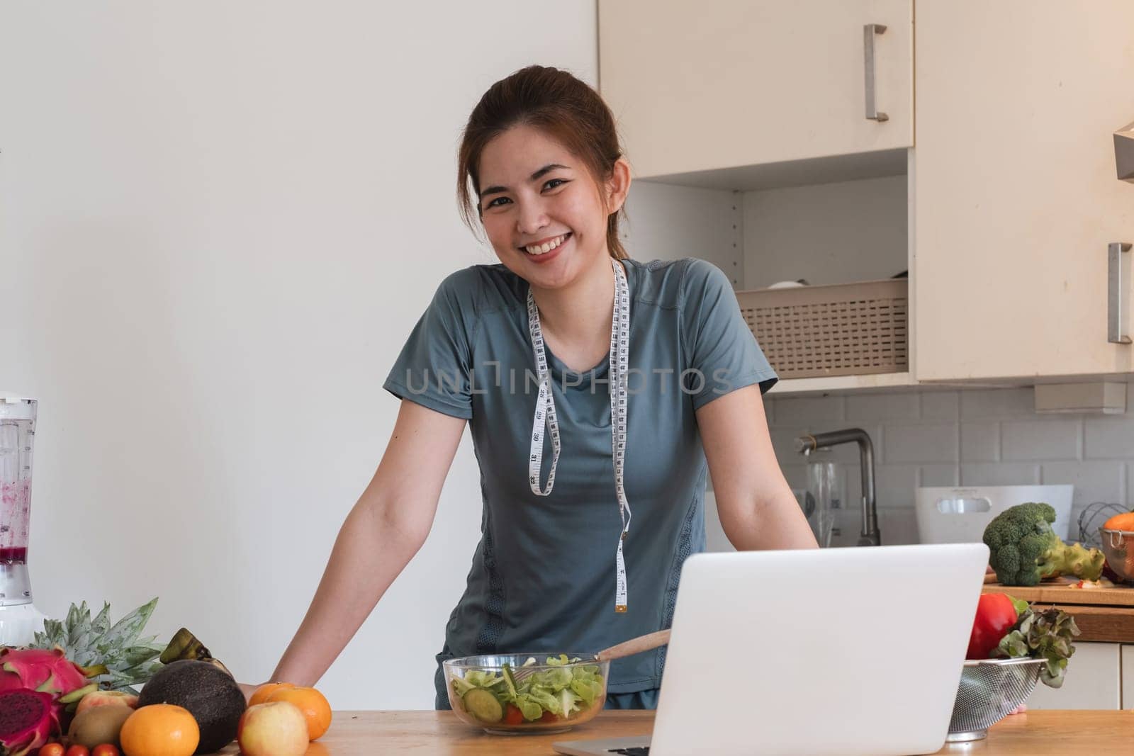 A woman is smiling and standing in front of a table with a laptop and a bowl of salad