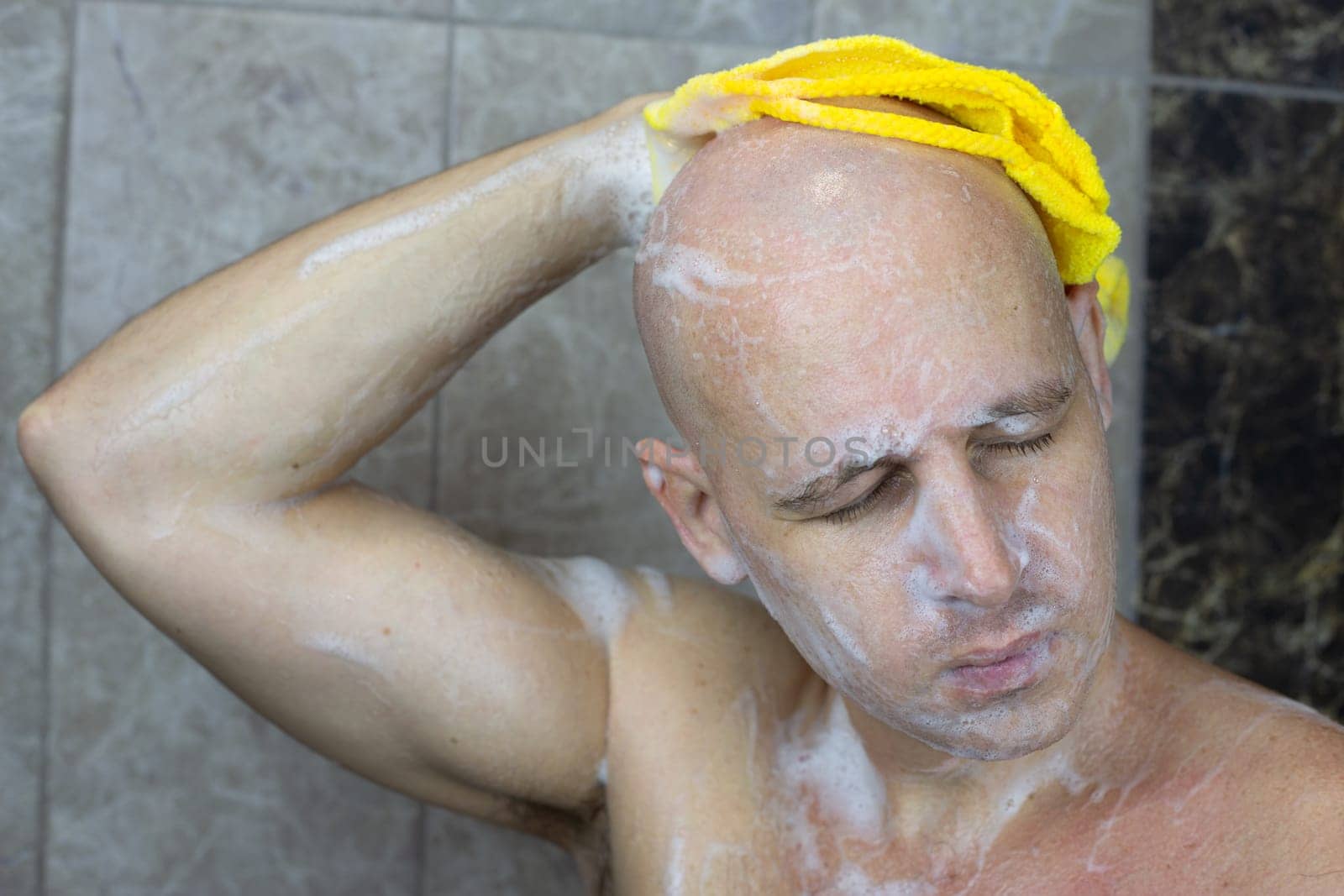 Man with closed eyes in shower washes his bald hairless head with washcloth and foam