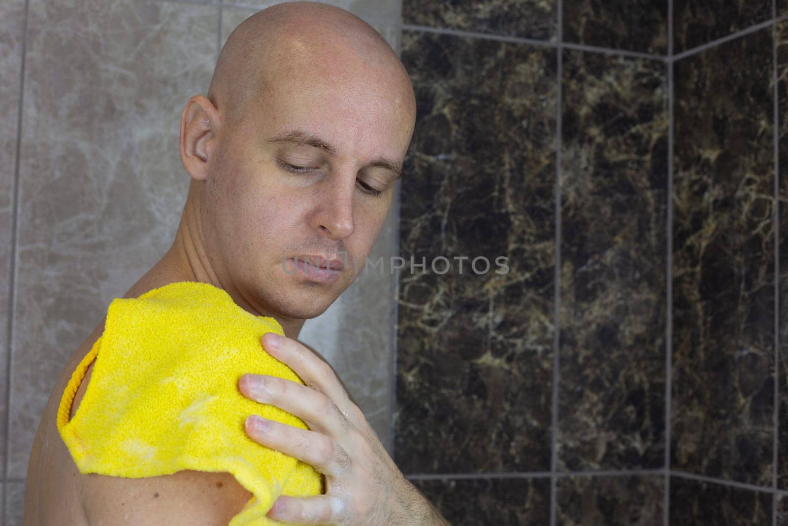 Man without hair washes himself in shower with washcloth, bald guy takes care of his skin in the shower with a special product with foam