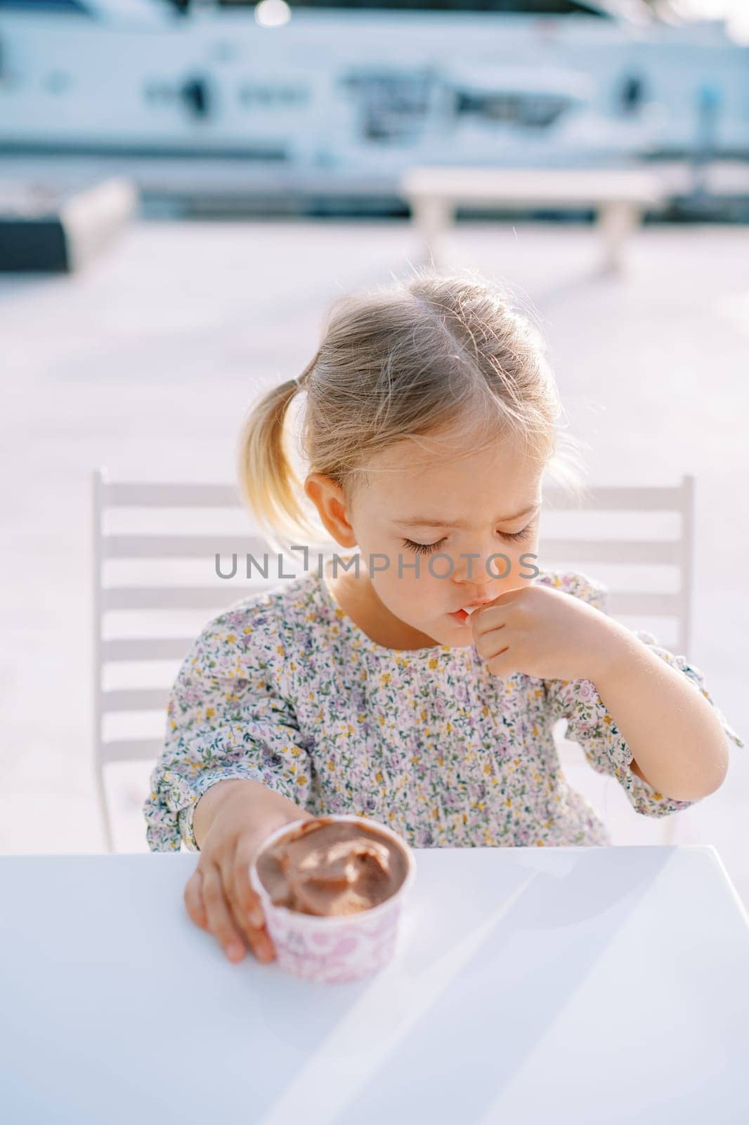 Little girl eats ice cream with a spoon from a cup while sitting at the table. High quality photo