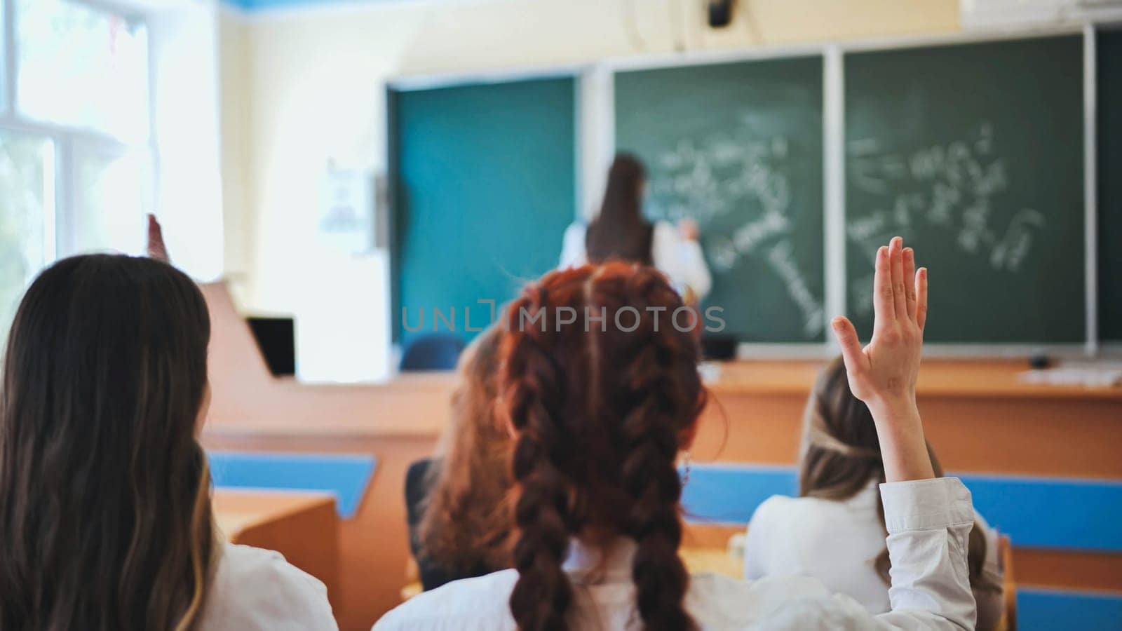 Intelligent group of young school children all raising their hands in the air to answer a question posed by the female teacher, view from behind