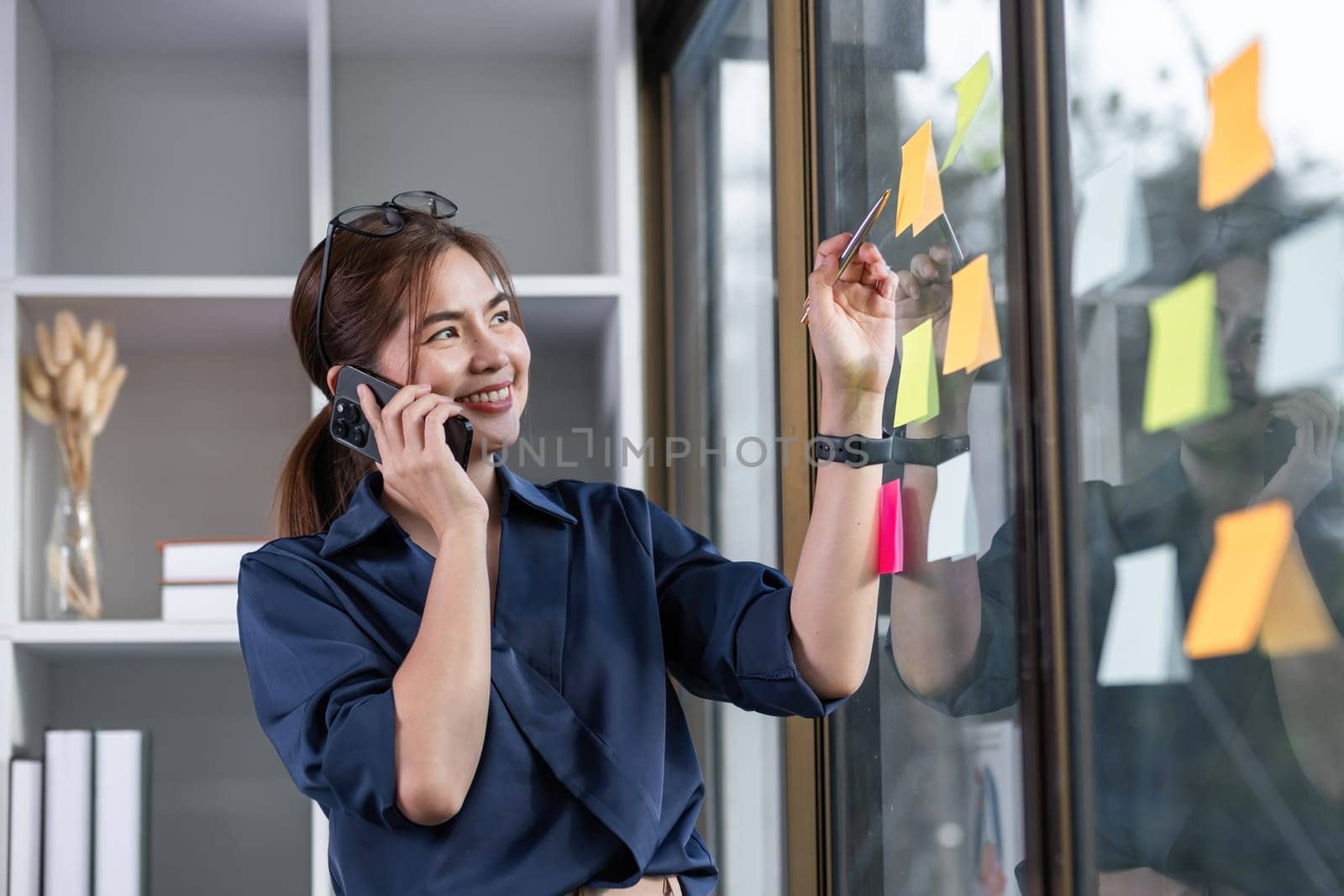 Asian businesswoman talking in a work meeting on the phone, taking notes on sticky notes on the wall in the office.