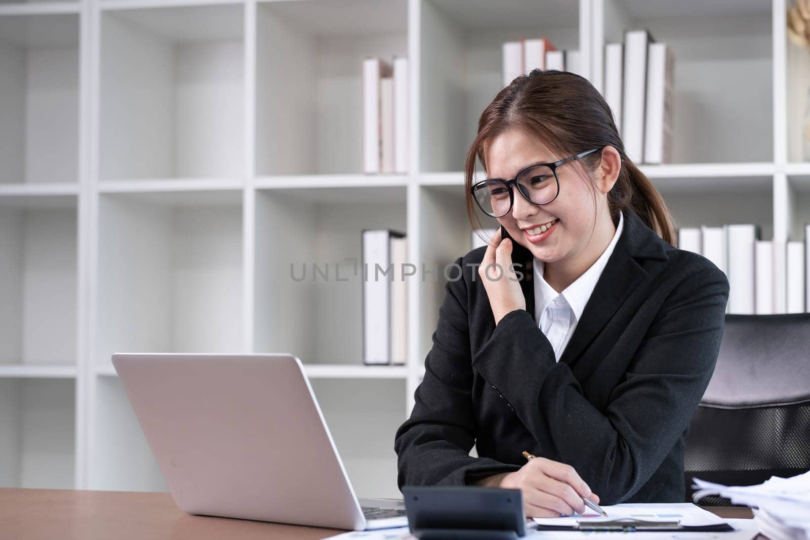 Asian female accountant is discussing business matters on the phone in the office with work documents on the table in the office..