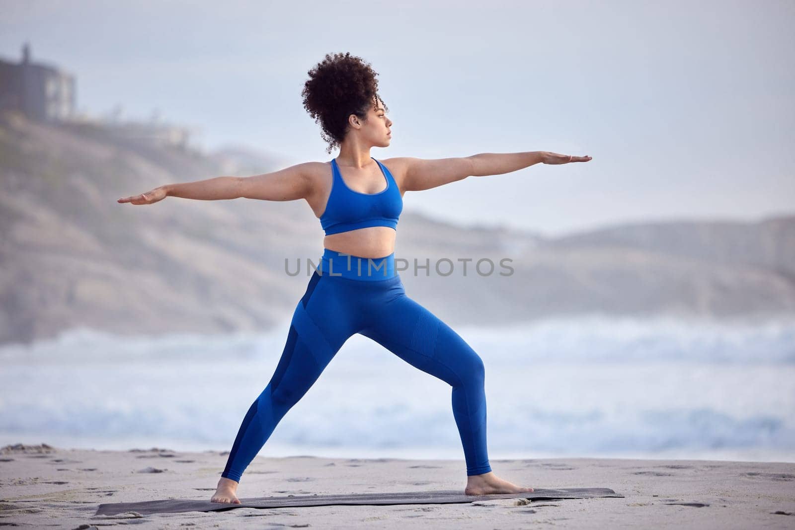 Black woman, yoga and fitness for wellness at beach with peaceful mind, warrior pose and body. African person, zen and pilates workout at sea with stretching, spirituality and balance with training.