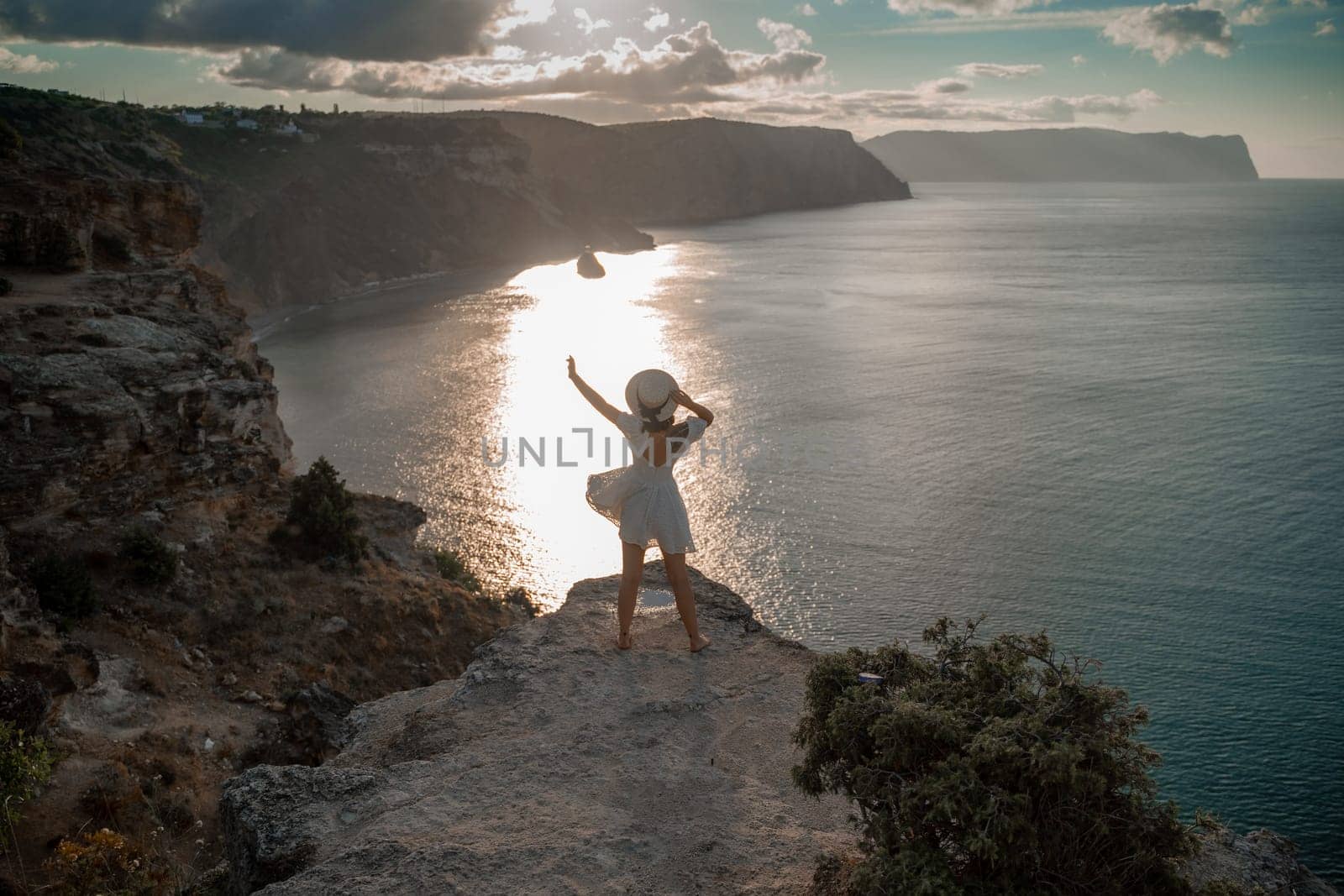 A woman stands on a cliff overlooking the ocean. She is wearing a white dress and a straw hat. The sky is cloudy and the sun is setting