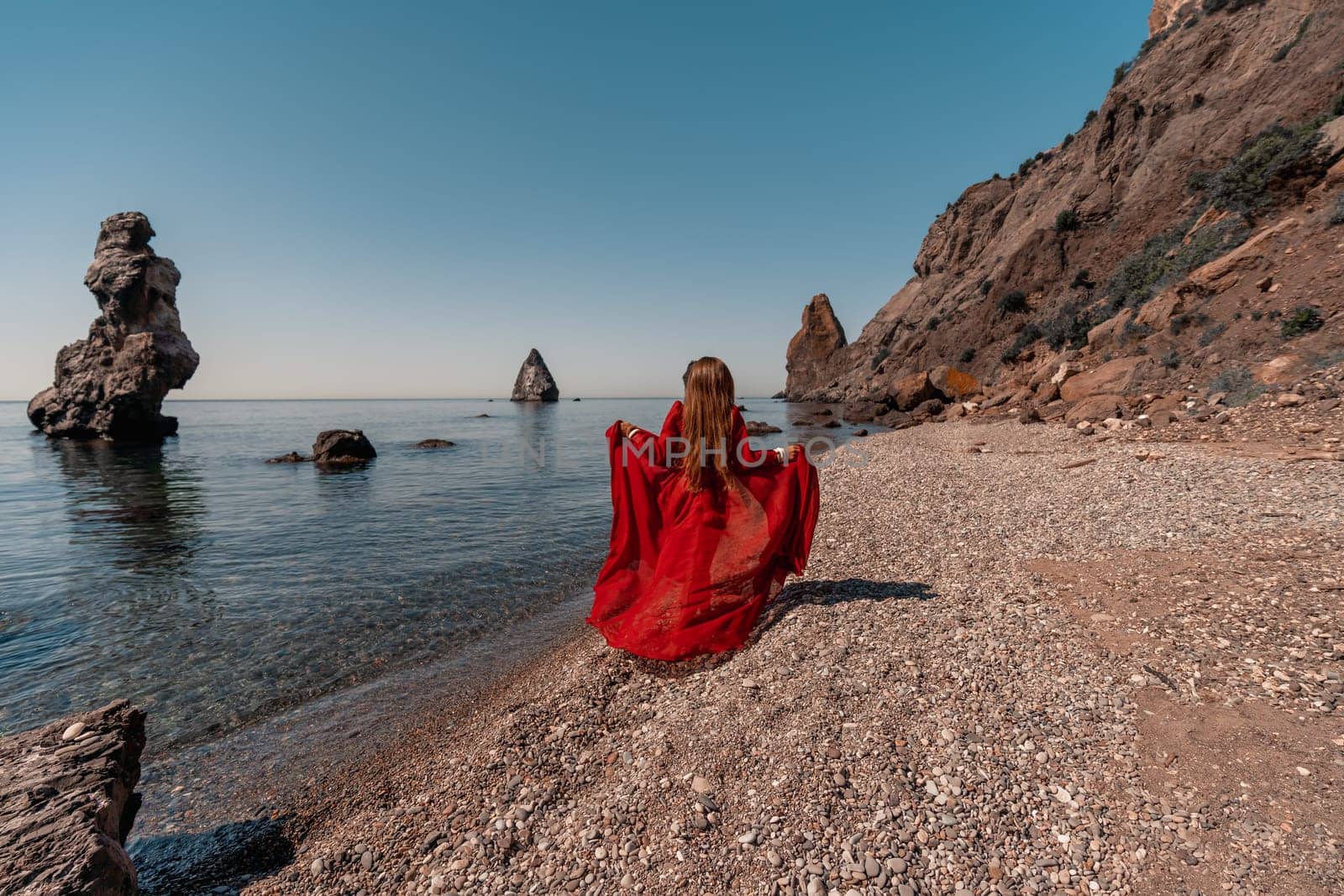 A woman in a red dress stands on a beach with a rocky shoreline in the background. The scene is serene and peaceful, with the woman's red dress contrasting against the natural elements of the beach