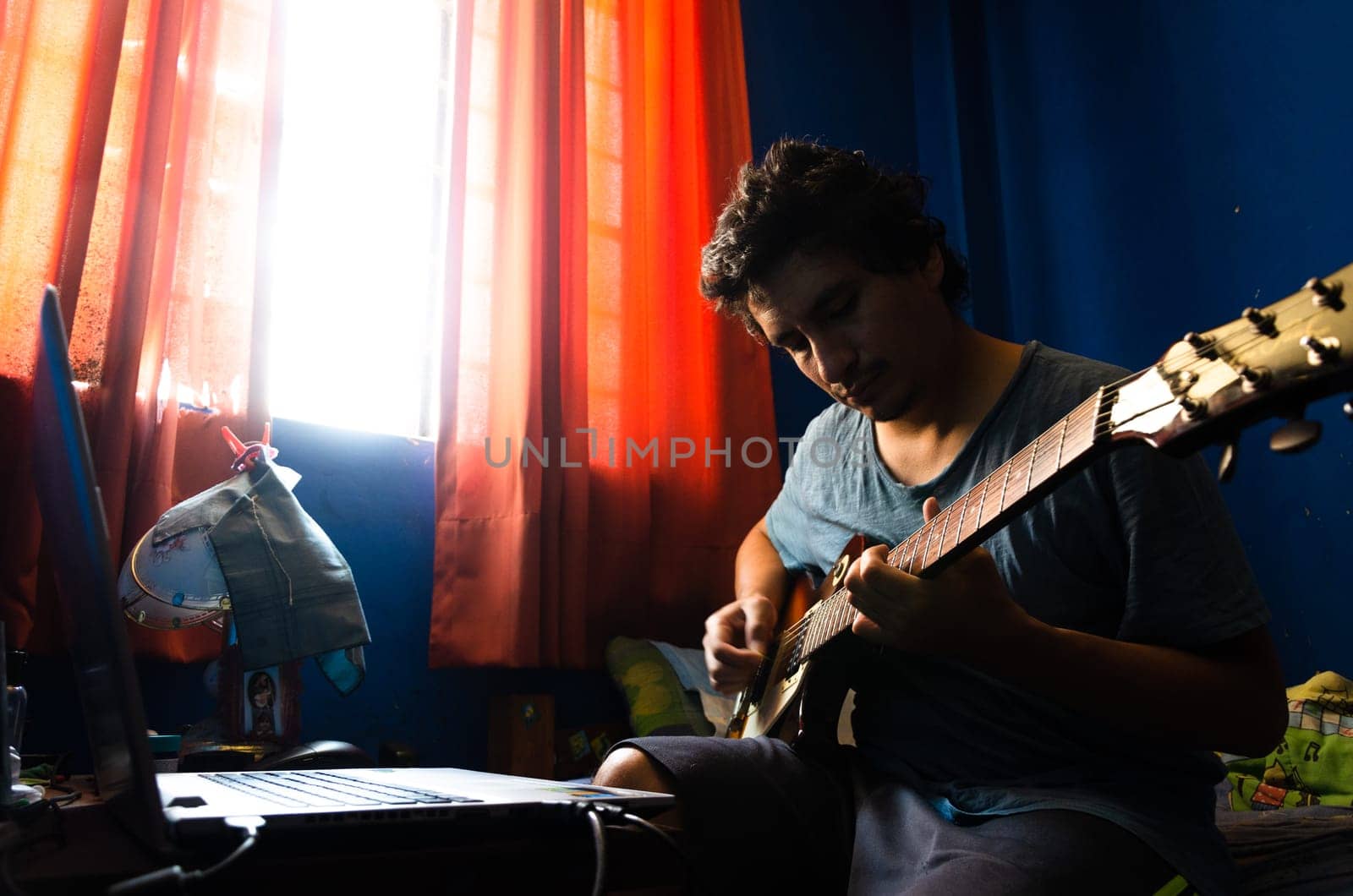 Casual young man with guitar and laptop in bed at home. by Peruphotoart
