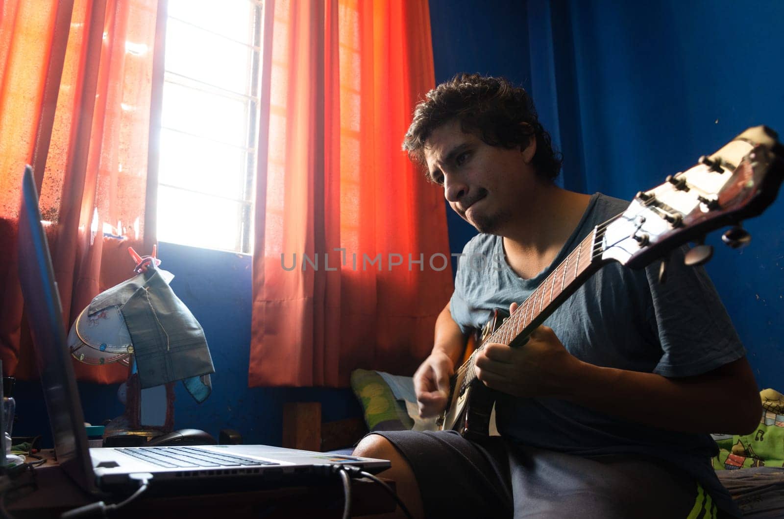 Casual young man with guitar and laptop in bed at home. by Peruphotoart