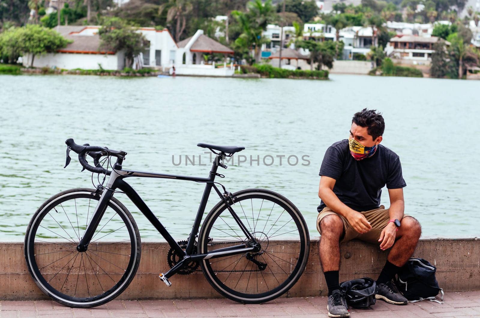 A young boy sitting on the edge of a lake with his bicycle with protective mask in times of pandemic. by Peruphotoart
