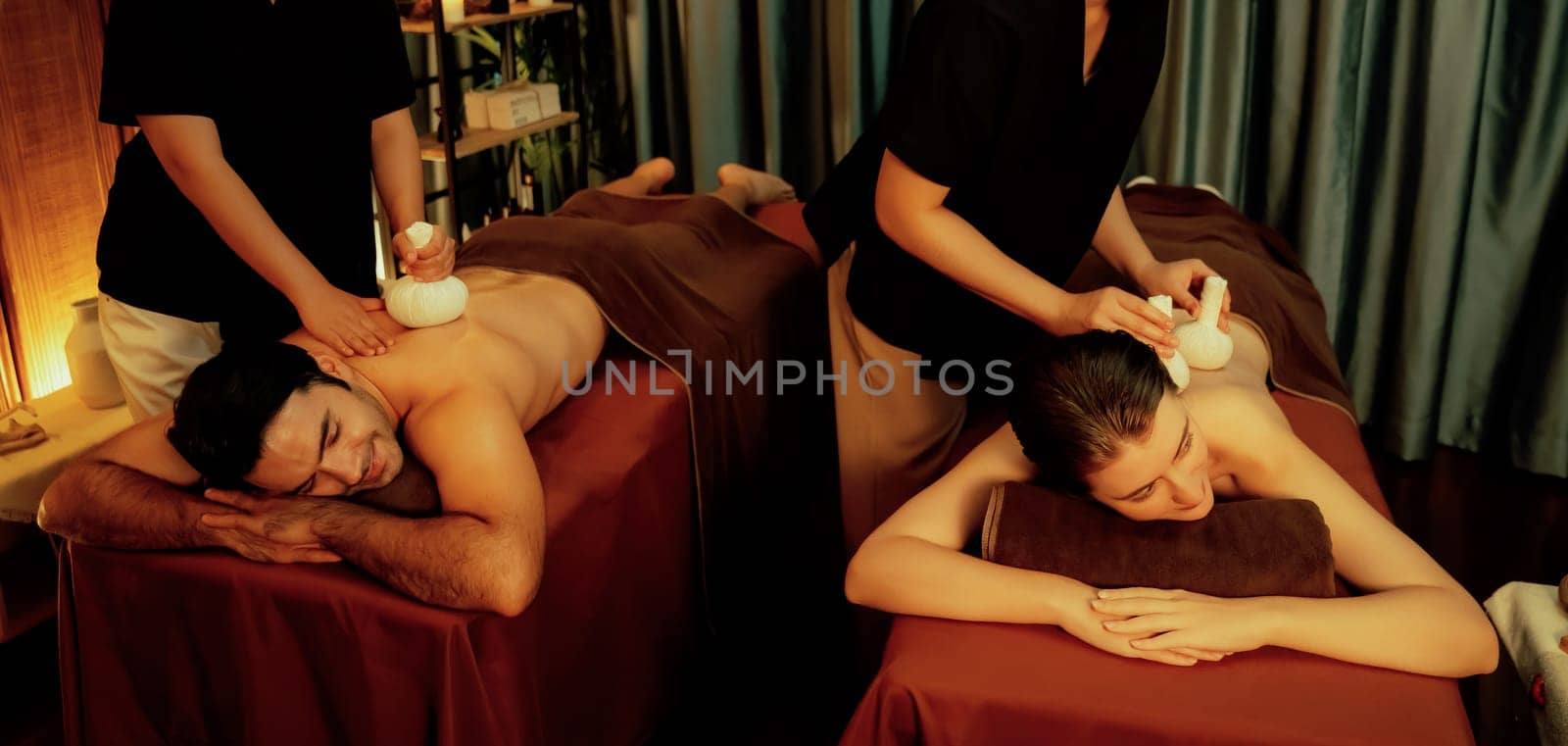 Hot herbal ball spa massage body treatment, masseur gently compresses herb bag on couple customer body. Serenity of aromatherapy recreation in warm lighting of candles at spa salon. Quiescent