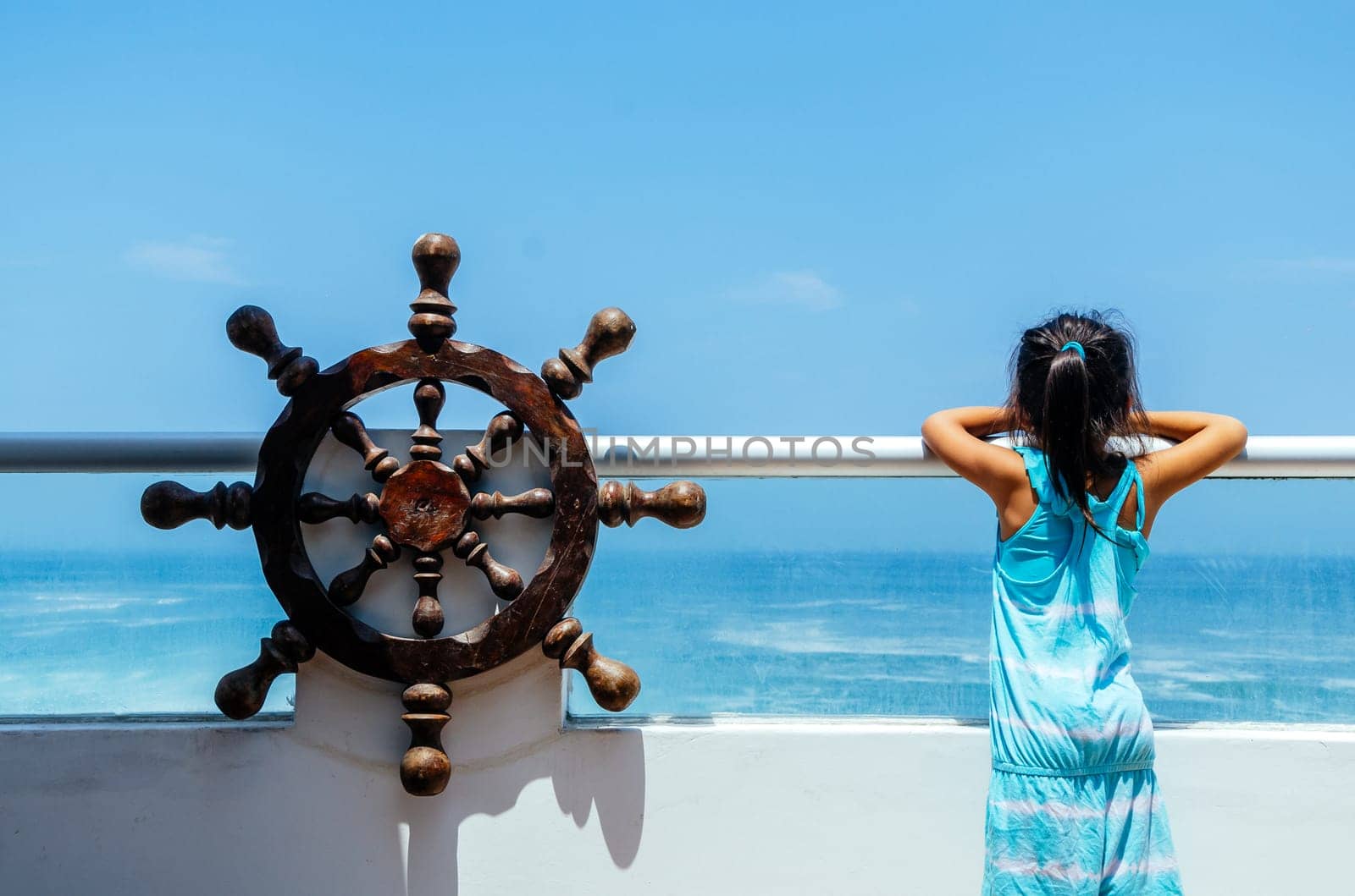 Little girl playing to be a sailor on a balcony with a boat rudder and the background of the sea. by Peruphotoart
