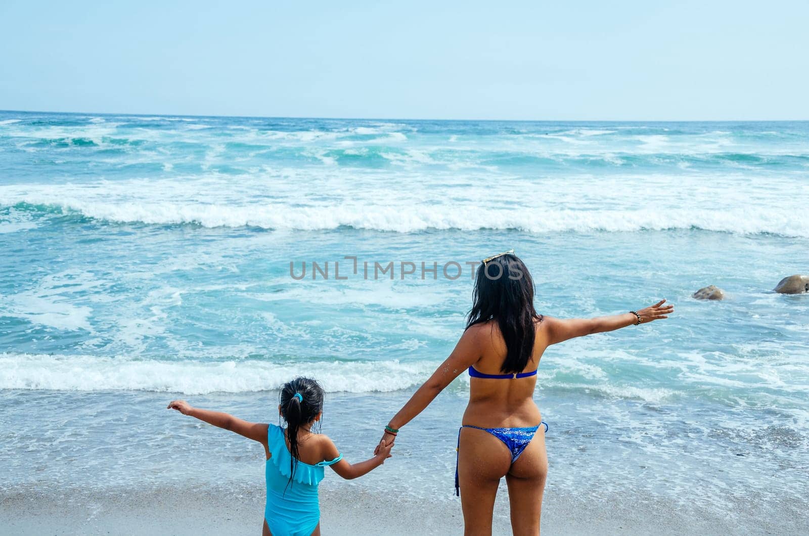 Action portrait of young mother with daughter on her back in front of the sea. Girls in swimsuits raising arms. by Peruphotoart