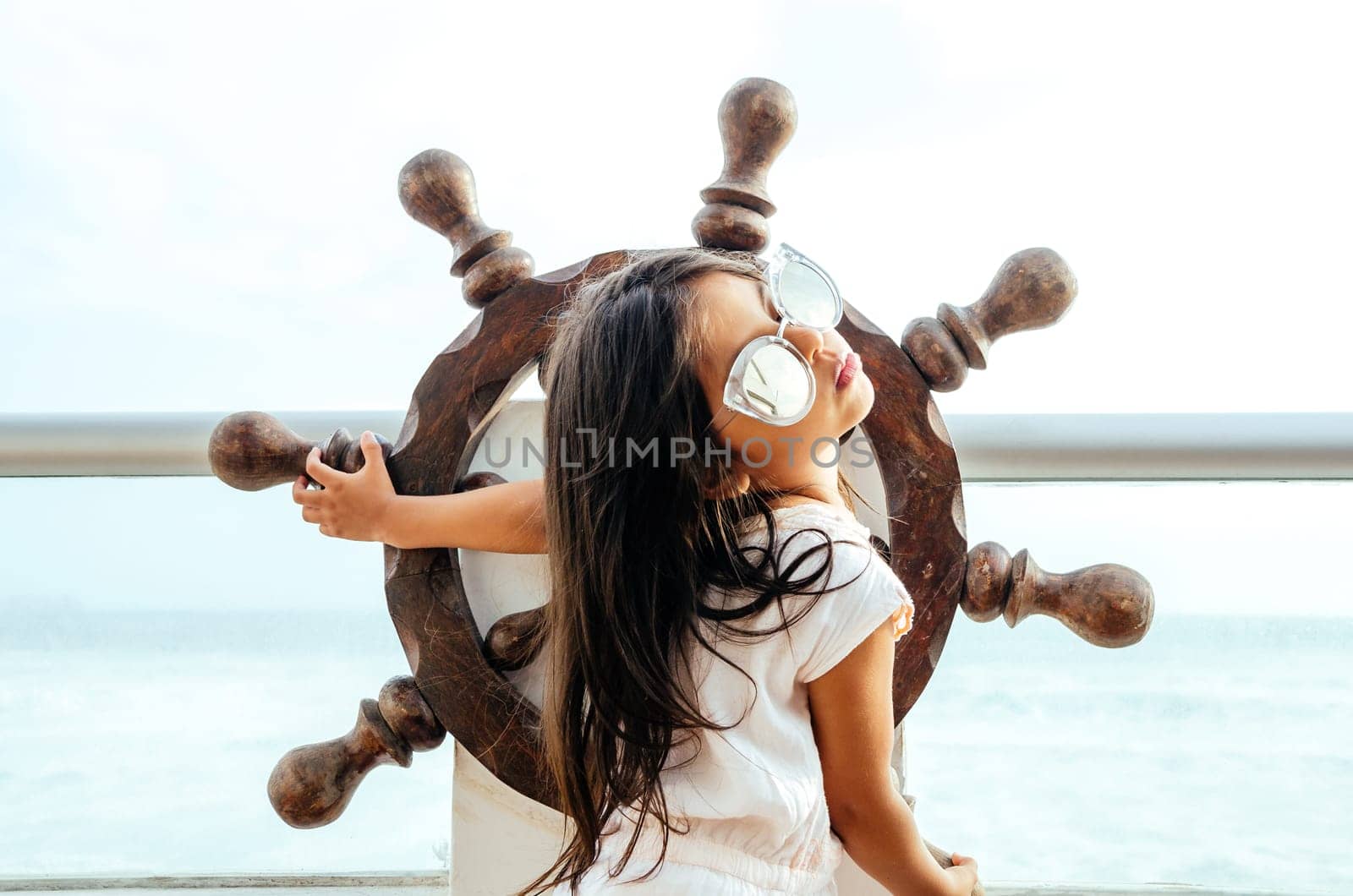 Little girl playing to be a sailor on a balcony with a boat rudder and the background of the sea