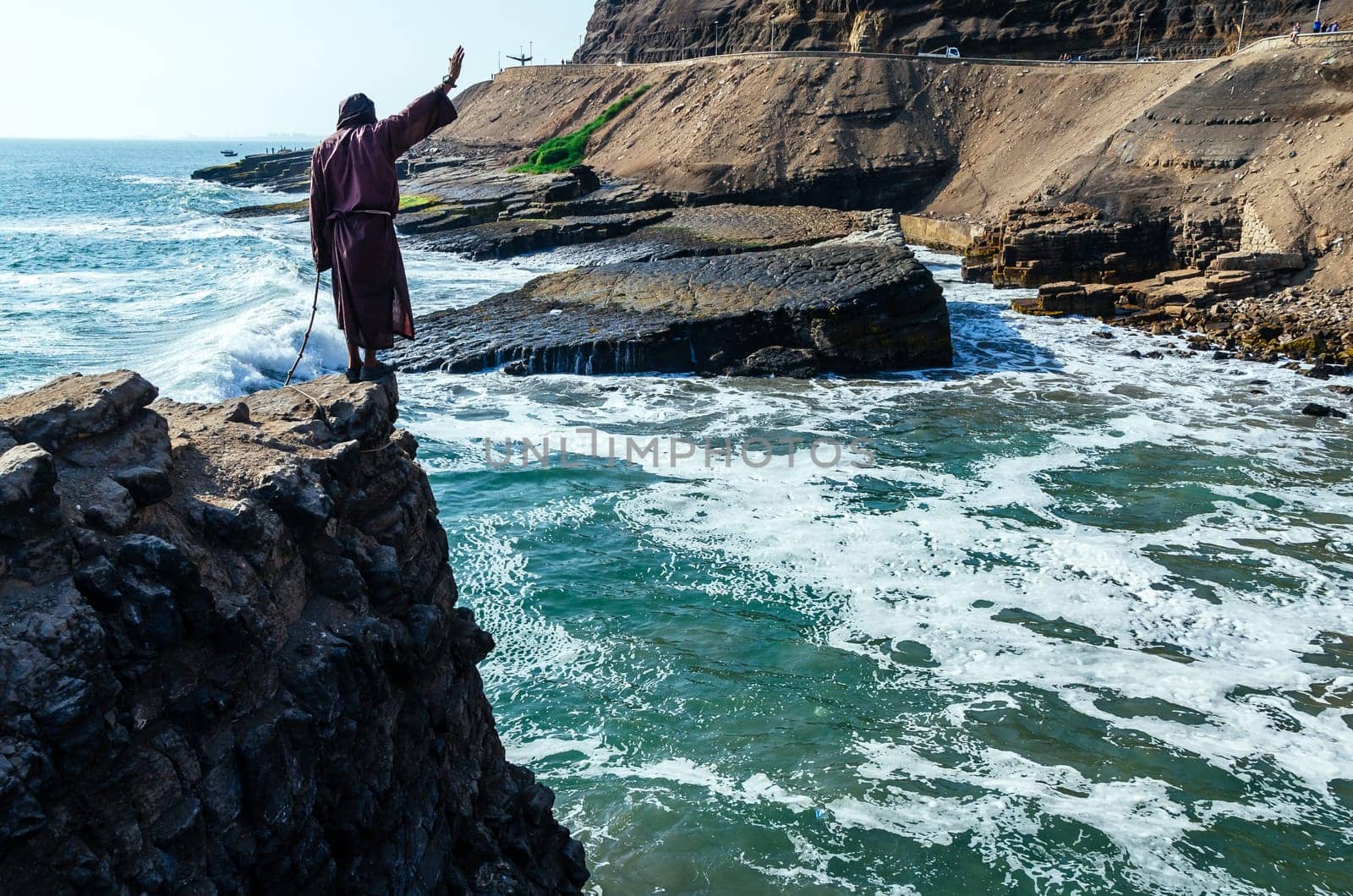 Man dressed as a friar throws himself off a cliff and head into the ocean at sunset. by Peruphotoart