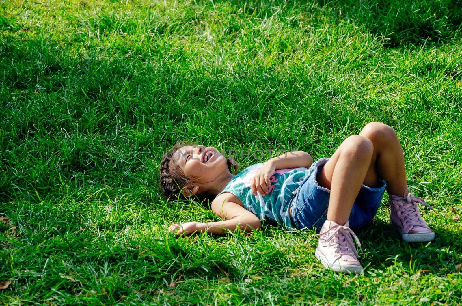 A little girl is smiling lying on the bright green grass. Portrait of a happy child on the grass in summer. The view from the top. Space for text
