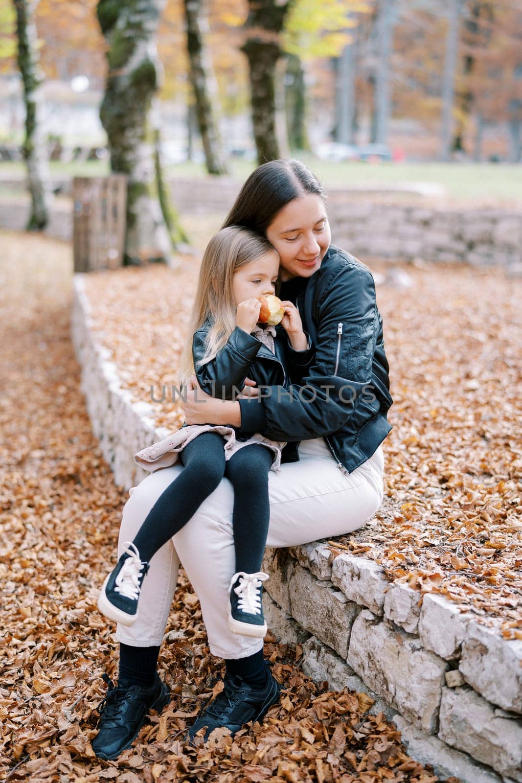 Mom hugs a little girl with an apple in her hands sitting on her lap on a low fence in the park. High quality photo