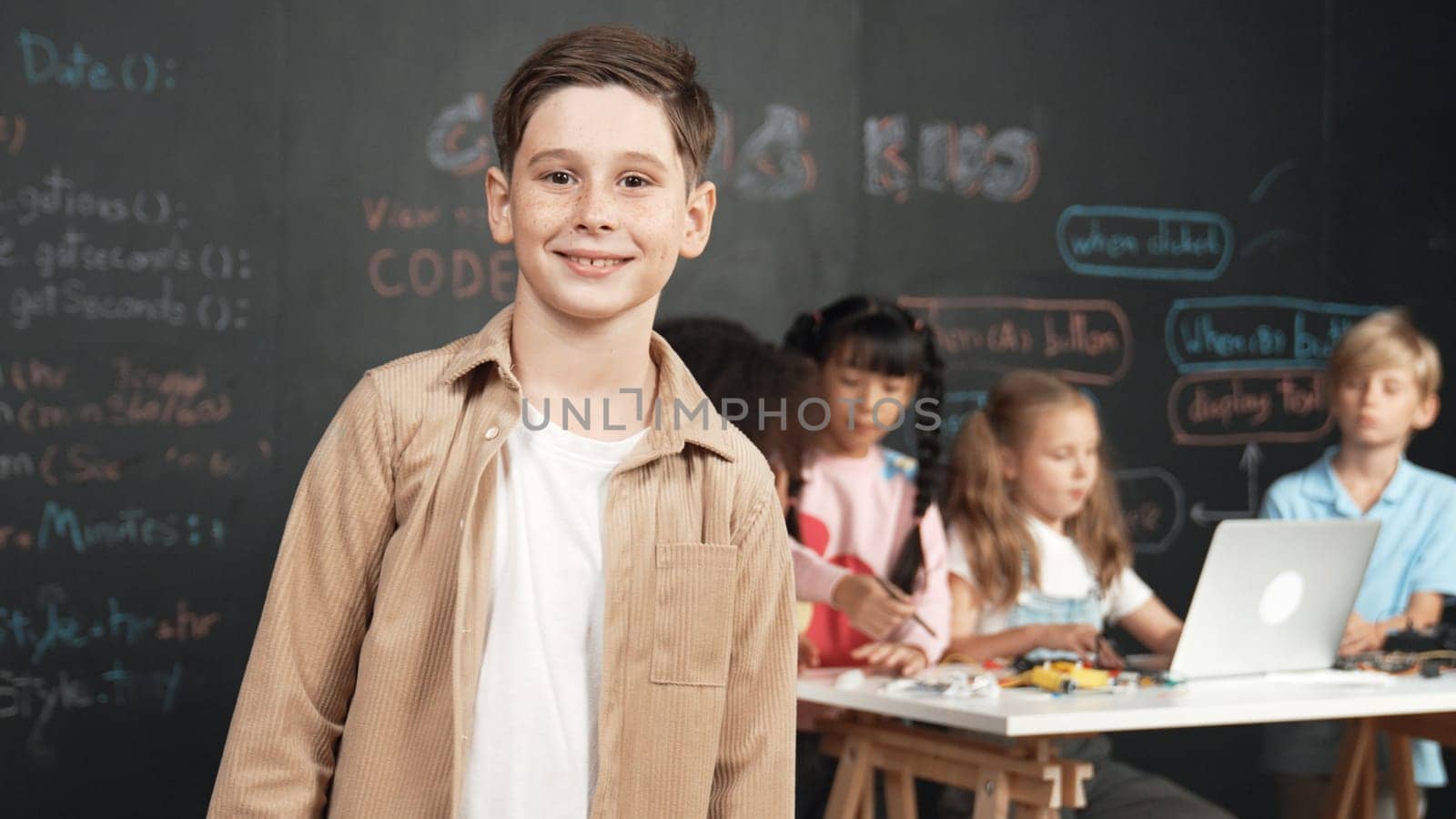 Smart boy smiling at camera while diverse friend working or learning engineering code or prompt in STEM technology classroom. Cute student standing at camera while children using laptop. Erudition.