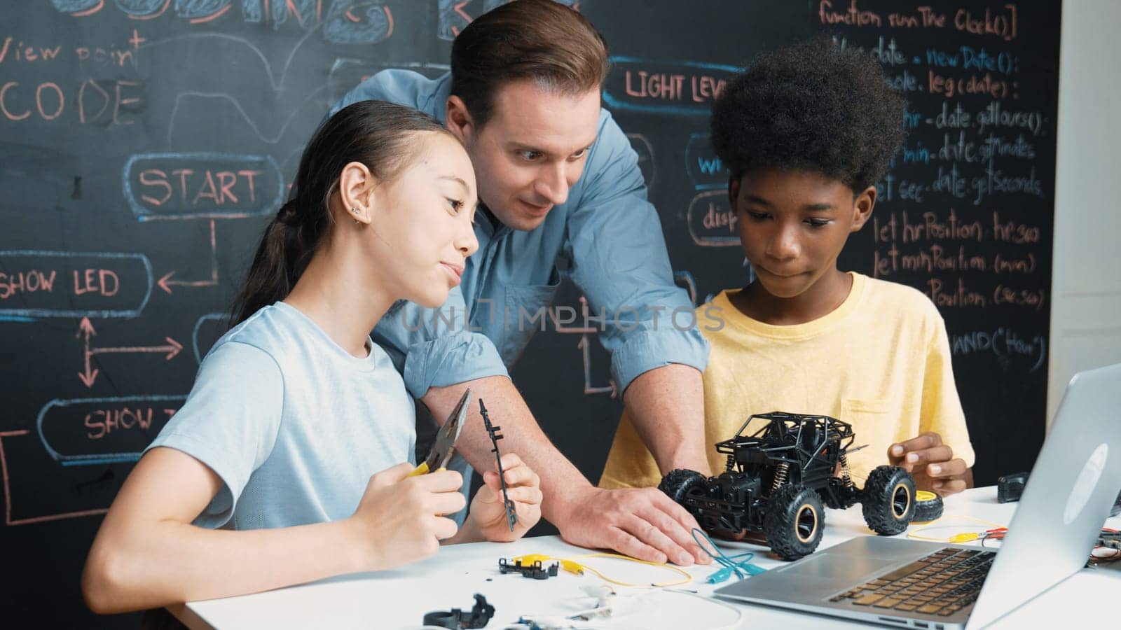Caucasian teacher looking at laptop screen while explain car model system at blackboard with coding or programing code written. Happy diverse student learning about robotic construction. Edification.