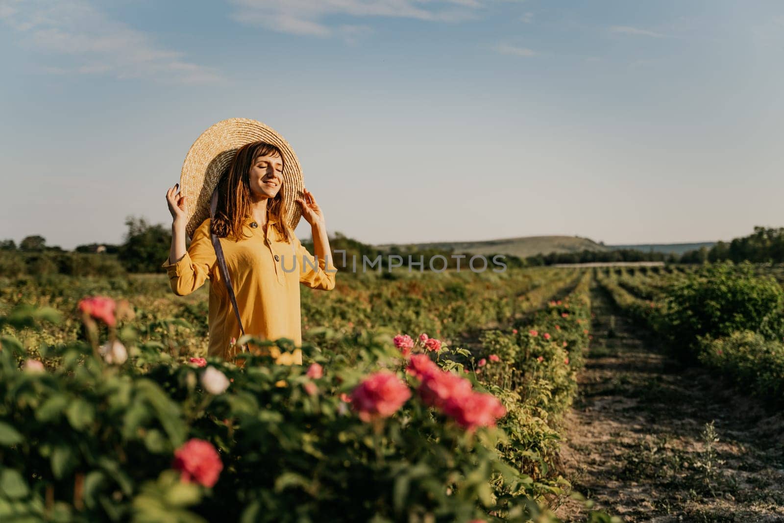 A woman wearing a straw hat stands in a field of pink flowers. She is smiling and she is enjoying the beautiful scenery. by Matiunina