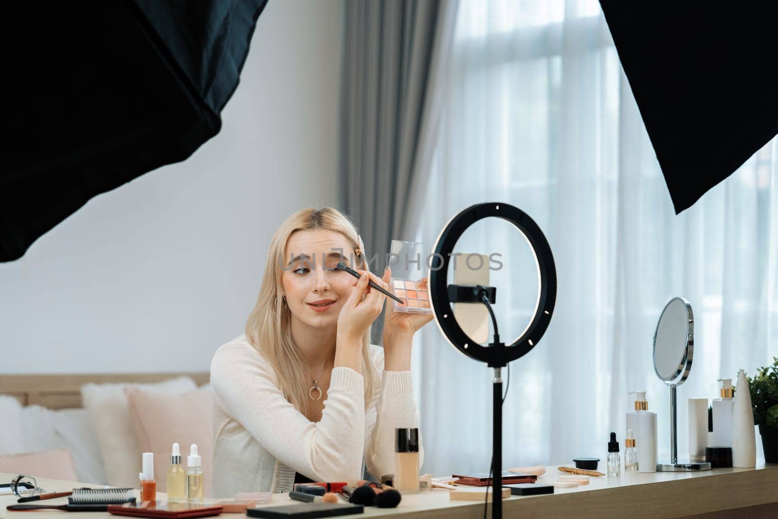 Young woman making beauty and cosmetic tutorial video content for social media. Beauty blogger using camera and light ring while showing how to apply eyeshadow to audience or follower. Blithe