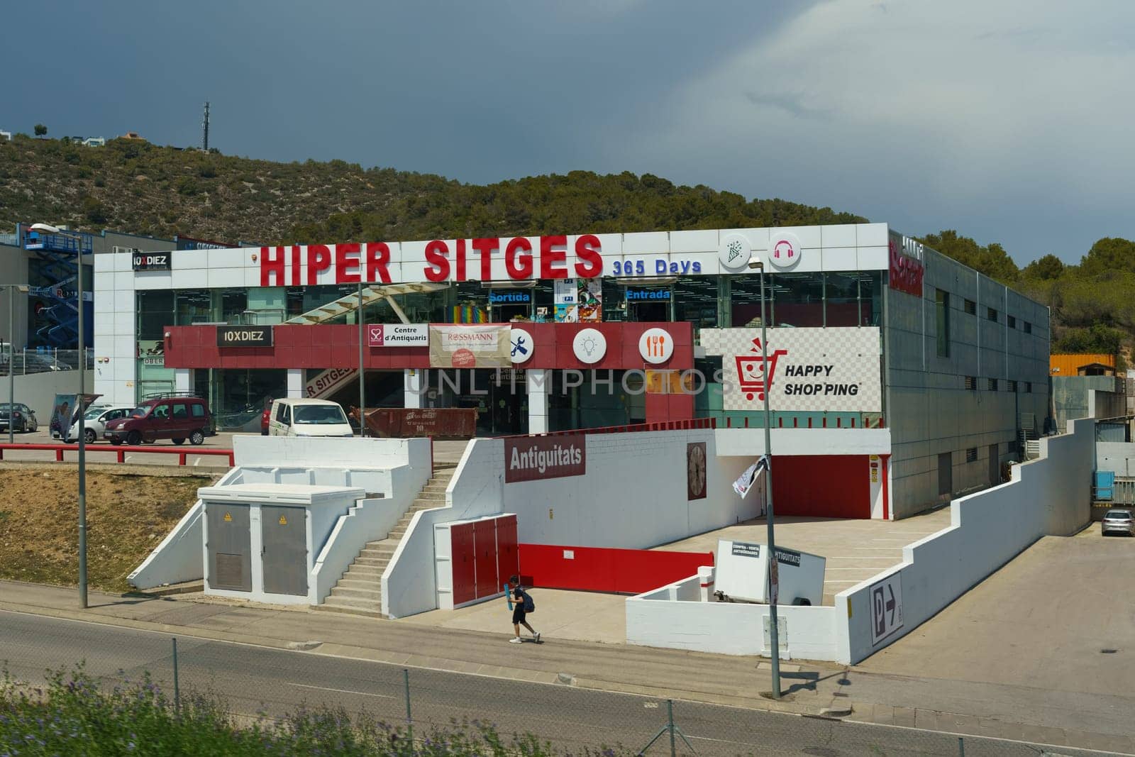 Barcelona, Spain - May 24, 2023: A contemporary two-story commercial building HIPER SITGES with bold signage, flanked by a road and green hills under a clear sky.