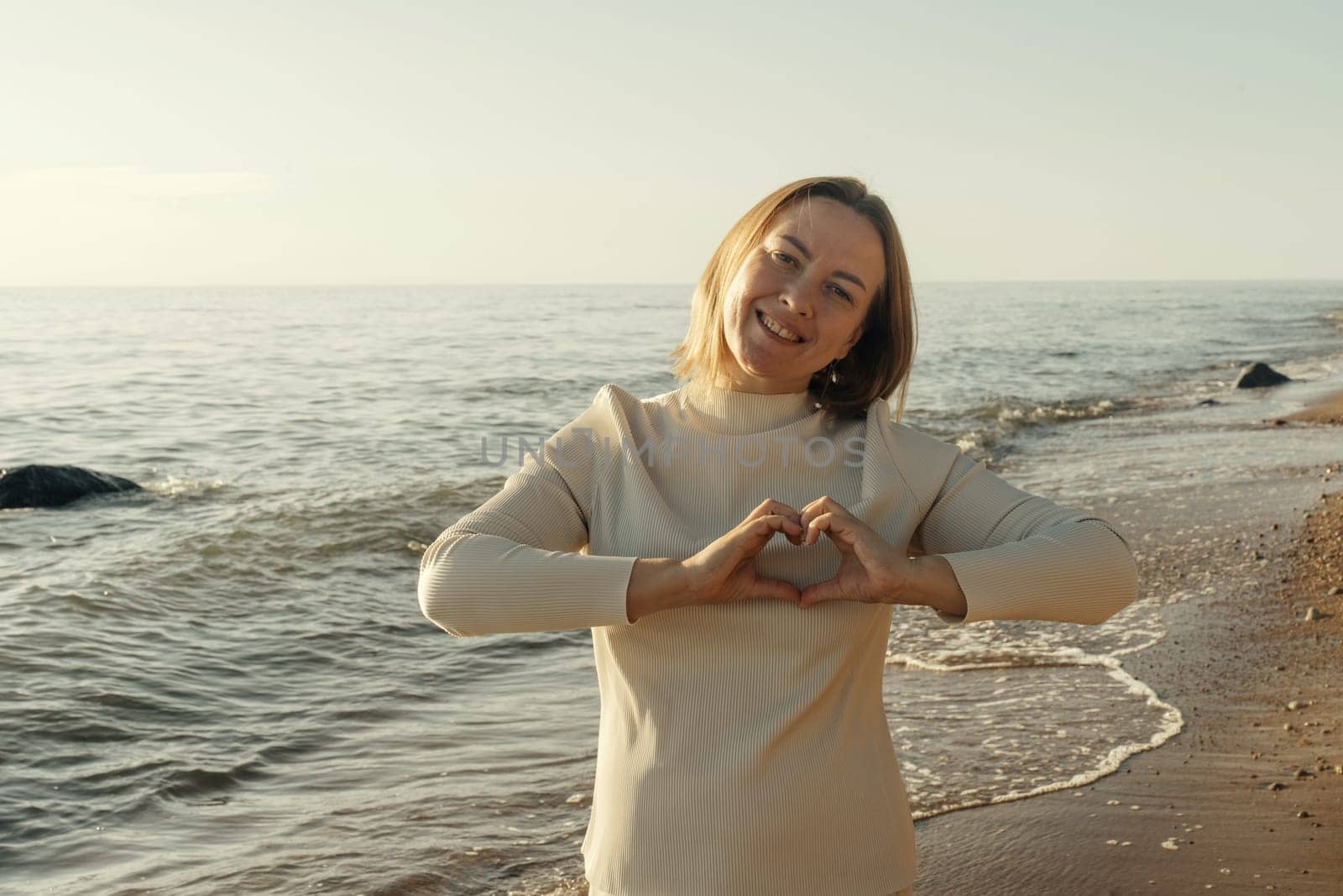 A woman is standing on a beach, forming a heart shape with her hands.