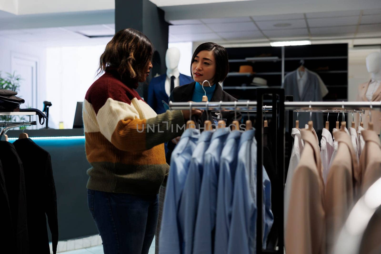 Clothing store woman asian assistant talking and consulting customer. Customer exploring rack with apparel, choosing formal outfit and getting help from boutique employee