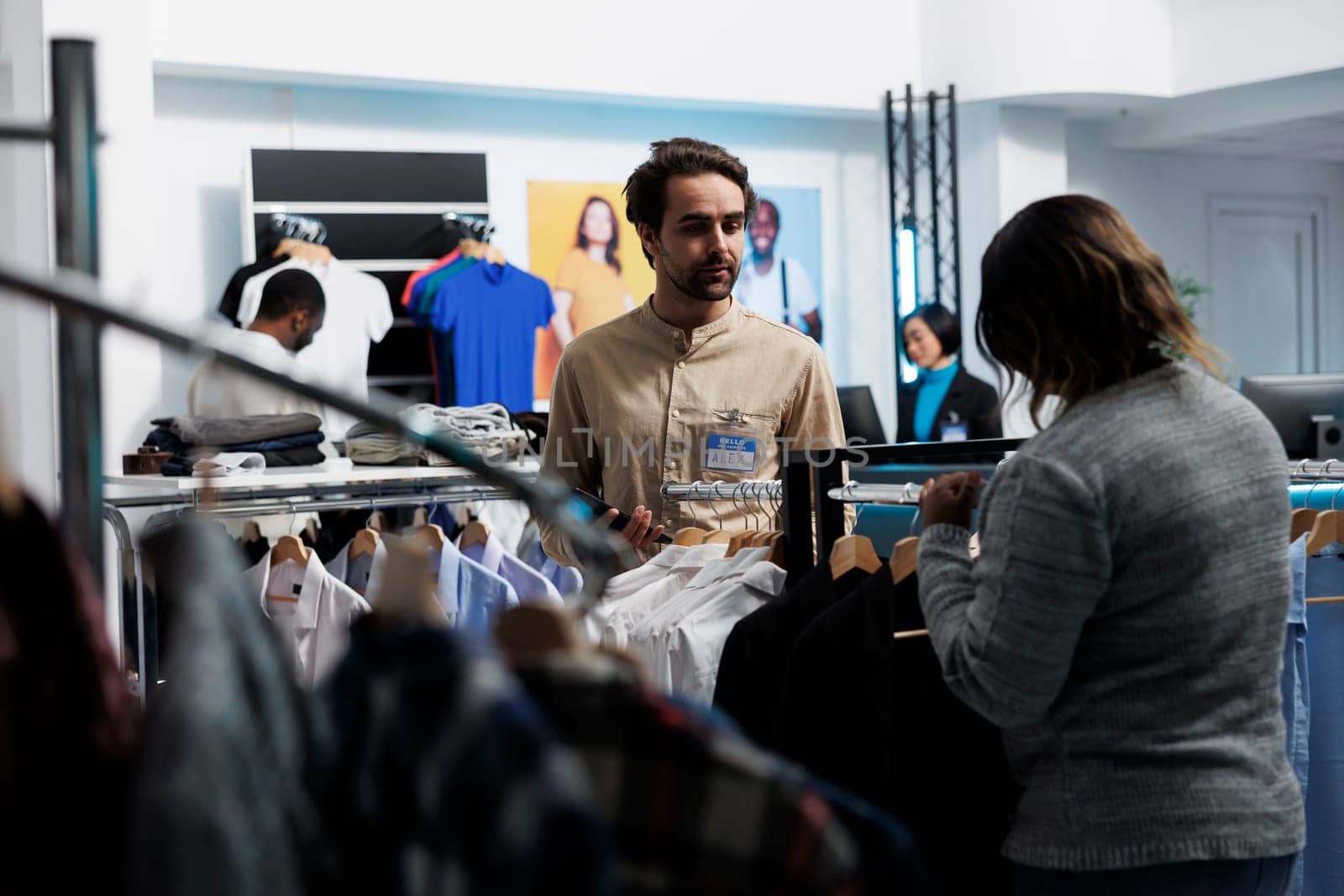 Clothing store consultant guiding customer through merchandise and using digital tablet. African american woman browsing apparel rack and talking with employee while shopping in mall
