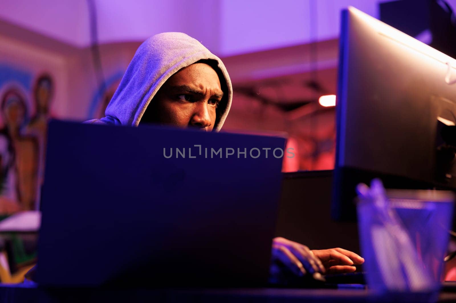 Focused african american scammer cracking password to gain access to system. Concentrated hacker in hood hacking database and breaching data in dark hideout place with neon light
