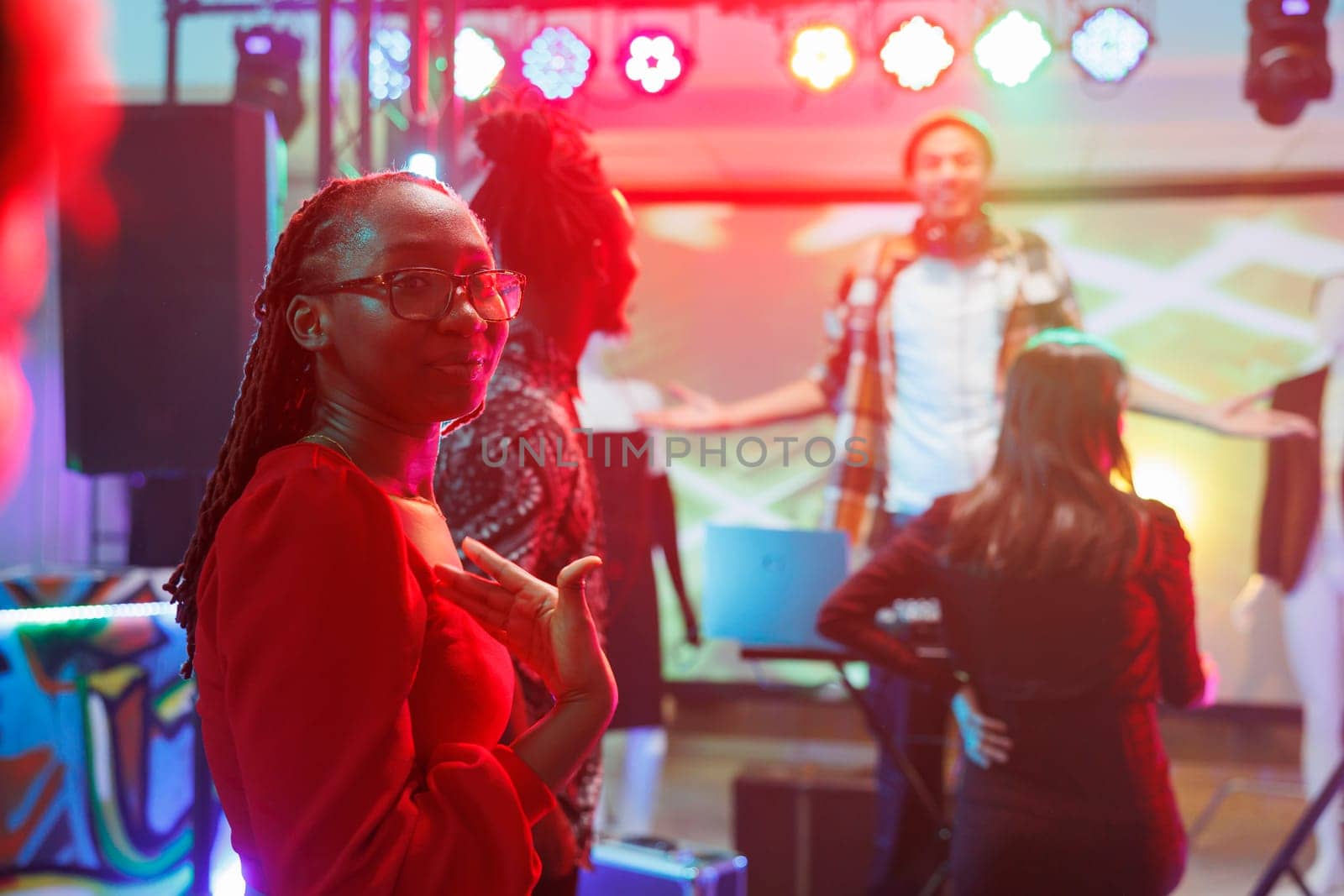 African american woman standing on dancefloor showing with hand at herself in nightclub. Young clubber having fun, relaxing and partying in club with spotlights, enjoying nightlife