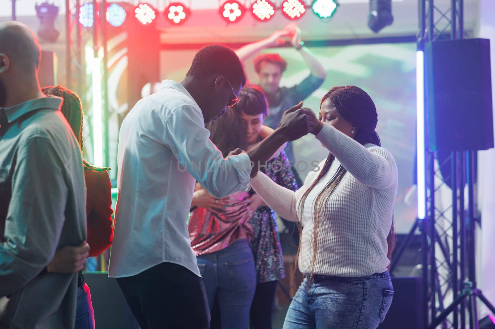 Young man and woman friends holding hands while dancing at nightclub party. African american couple clubbing and moving on dancefloor while attending discotheque gathering event