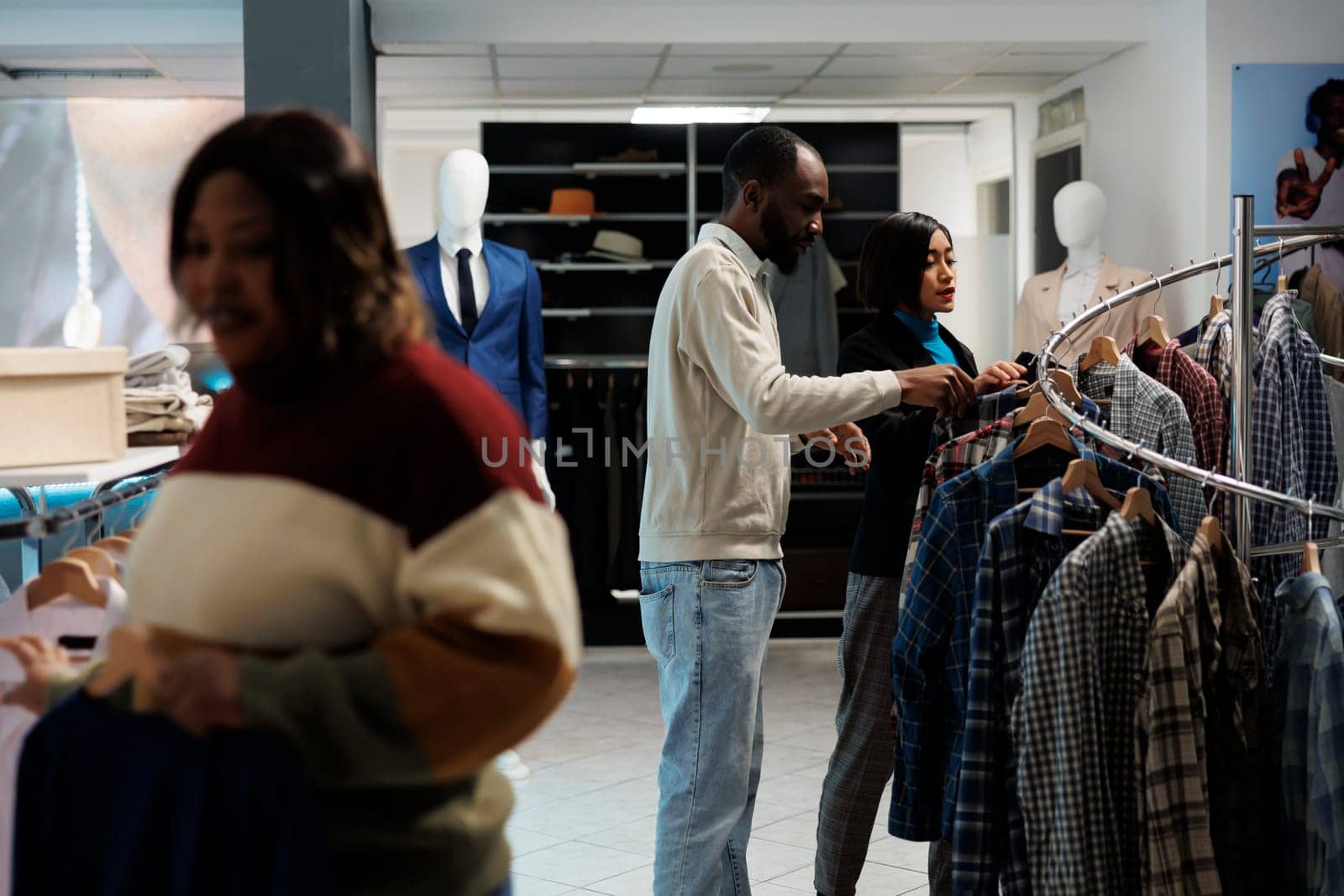 Clothing store assistant and buyer browsing apparel rack together while choosing casual trendy outfit. Boutique asian woman worker offering client advice in selecting shirt size