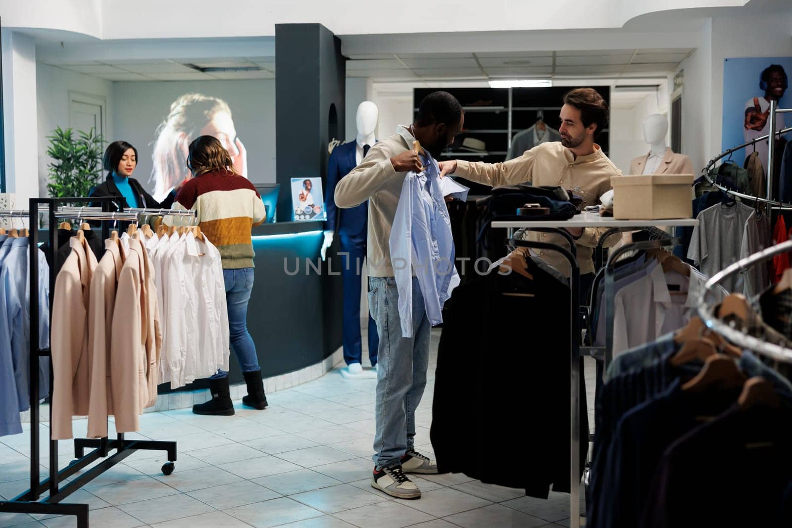 Clothing store customer trying on shirt and getting advice from boutique employee. African american man holding apparel to body while choosing size with fashion outlet assistant