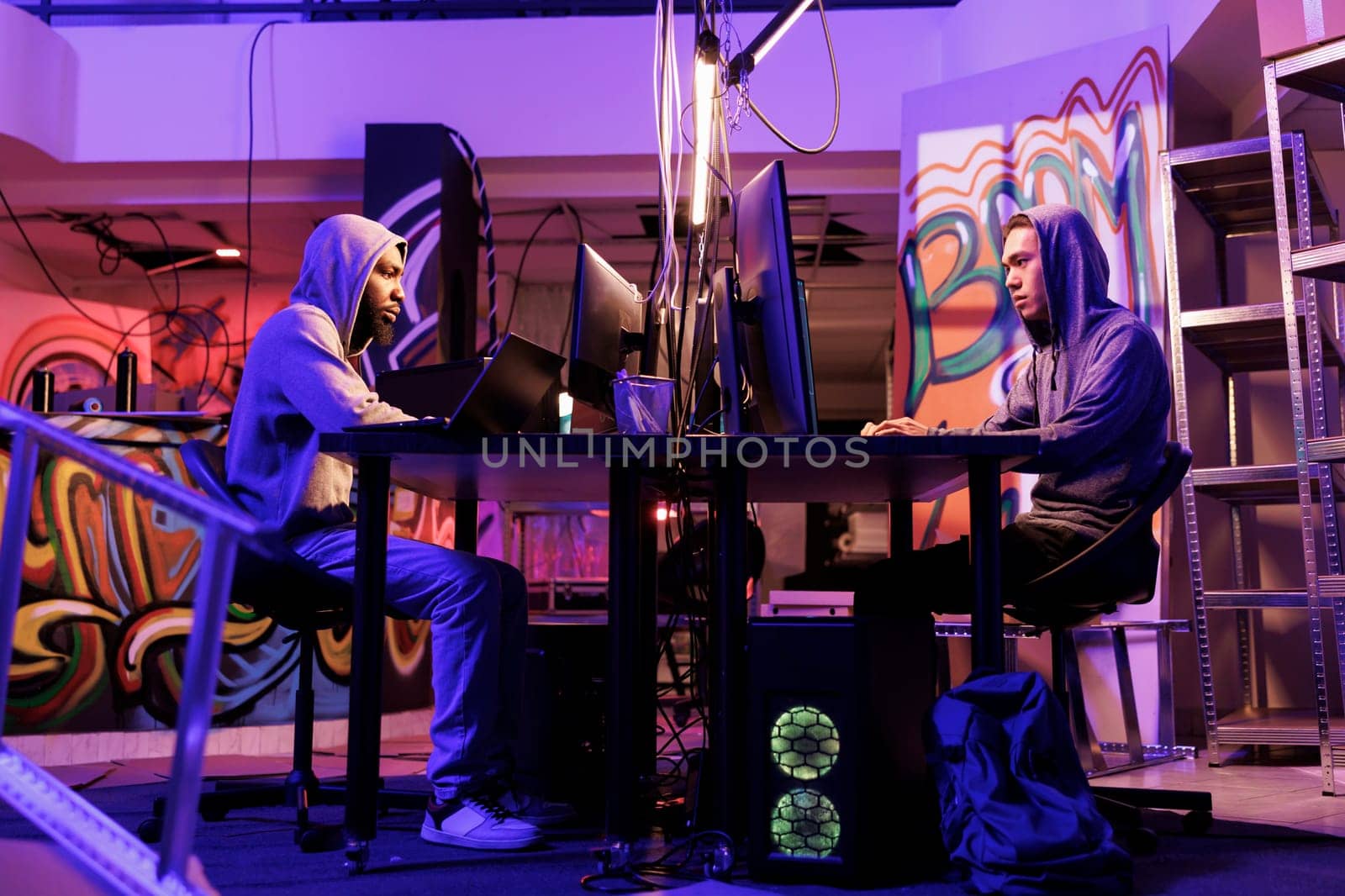 Hackers writing malicious code while working together at hideout workspace. African american and asian criminals programming internet virus to infect computer systems in abandoned warehouse