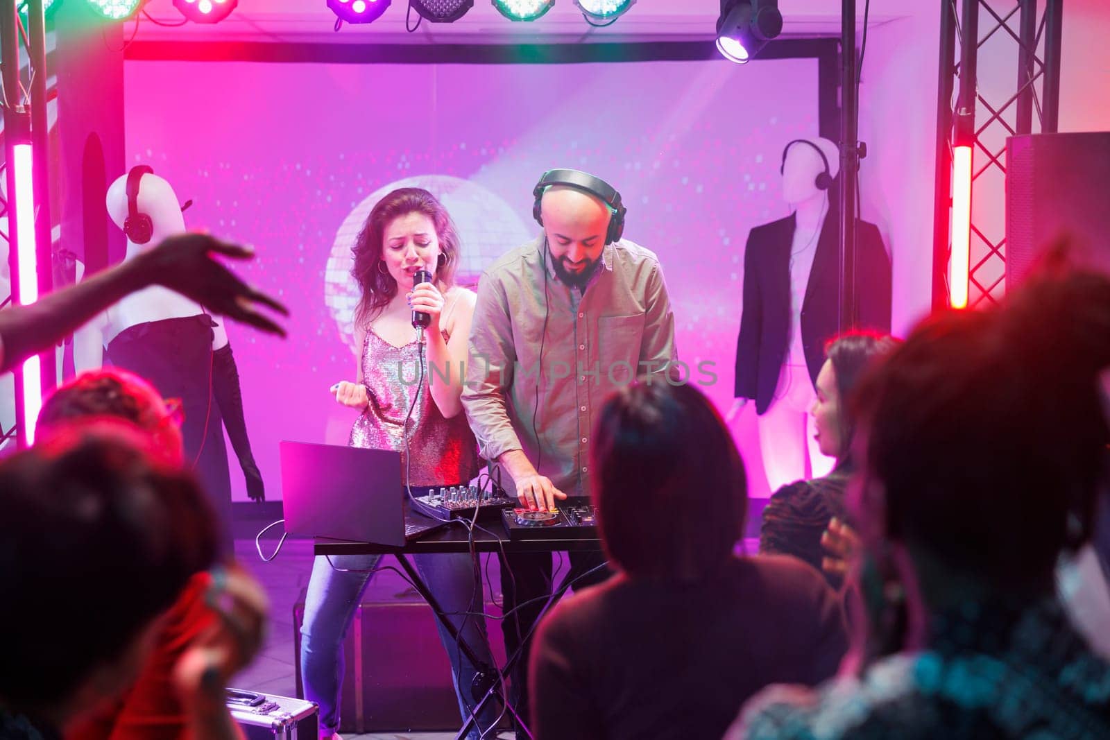 Woman singing and man dj mixing tracks while people partying on dancefloor in nightclub. People dancing and enjoying electronic music band show on stage with spotlights in club