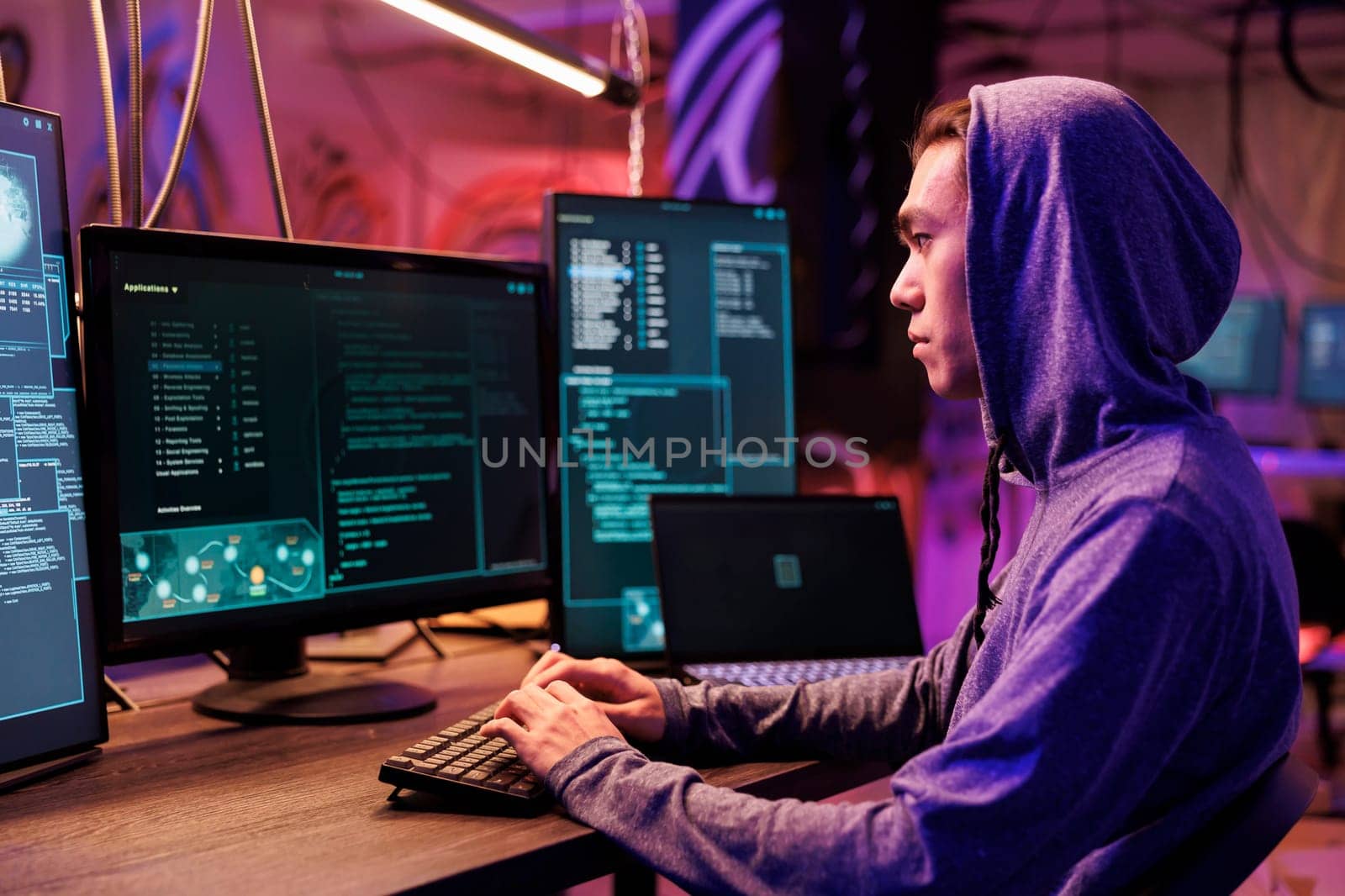 Asian hacker typing on keyboard and writing spyware code on computer. Internet criminal in hood hacking online server and planning phishing cyberattack in room at night time