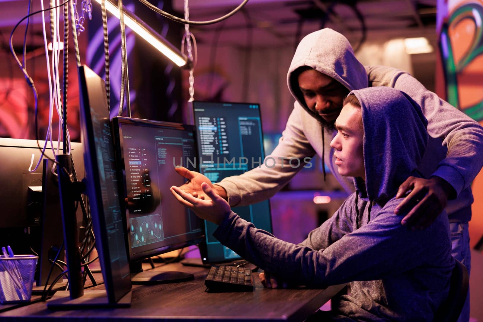 Two hackers collaborating while breaching data and coding malicious software on computer. Diverse criminals working together while programming ransomware in abandoned warehouse