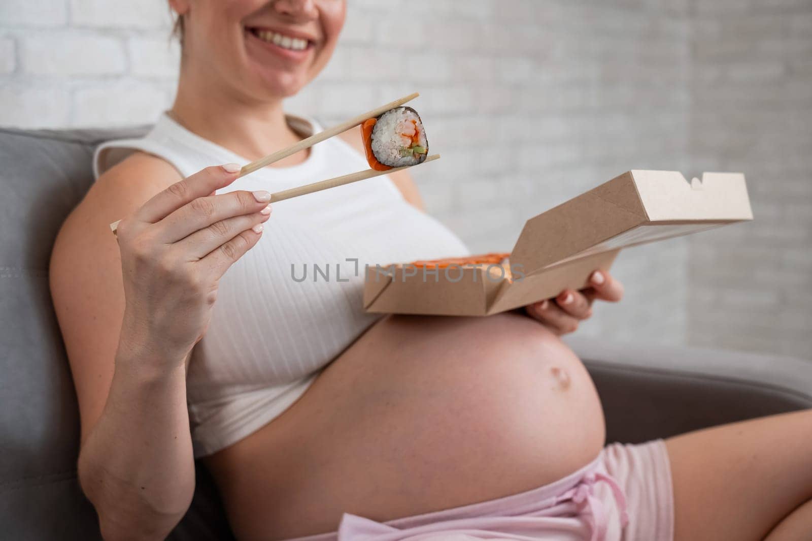 A pregnant woman sits on the sofa and eats rolls from a box. Food delivery