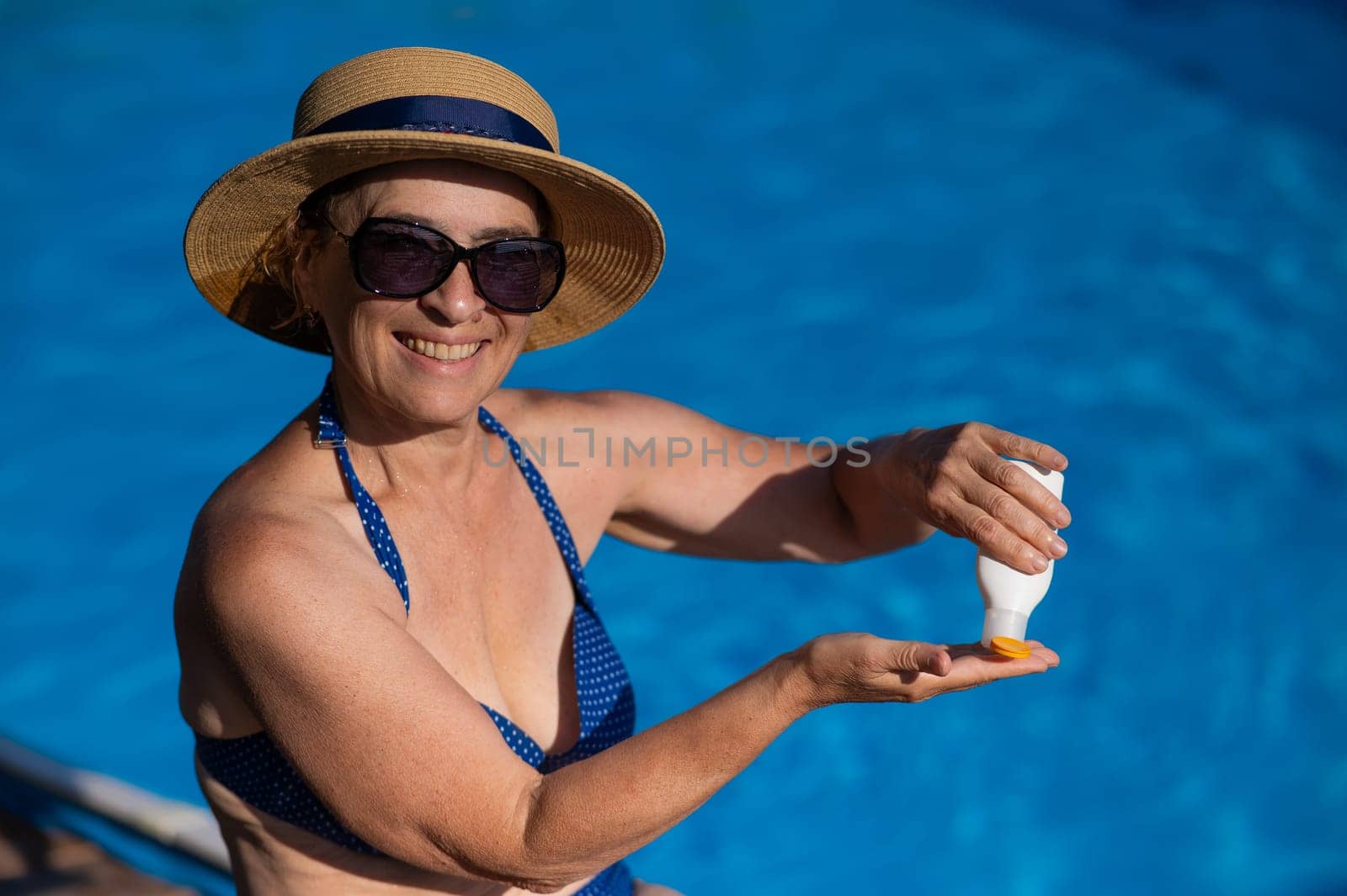Portrait of an old woman in a straw hat, sunglasses and a swimsuit applying sunscreen to her skin while relaxing by the pool