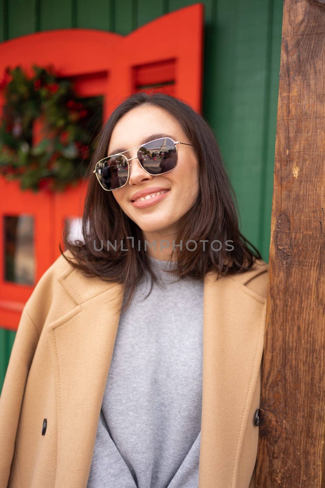 People, season and Christmas concept portrait of happy smiling middle age woman outdoors dressed stylish cream trench coat with Christmas festive decoration on background. Looking at camera