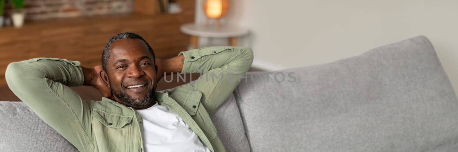 Cheerful middle aged african american man resting on sofa, enjoying free time alone, silence and comfort in cozy living room interior, close up, copy space. Relax, weekend break at home and lifestyle
