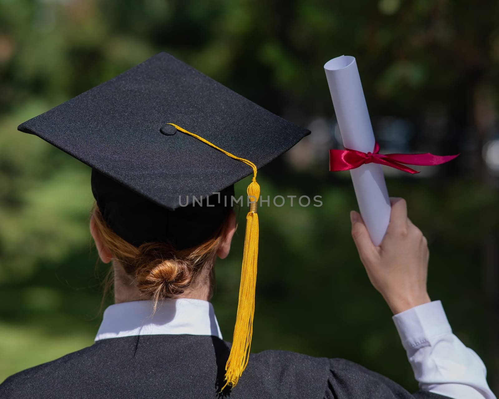 Rear view of a caucasian woman in a graduate gown holding a diploma outdoors