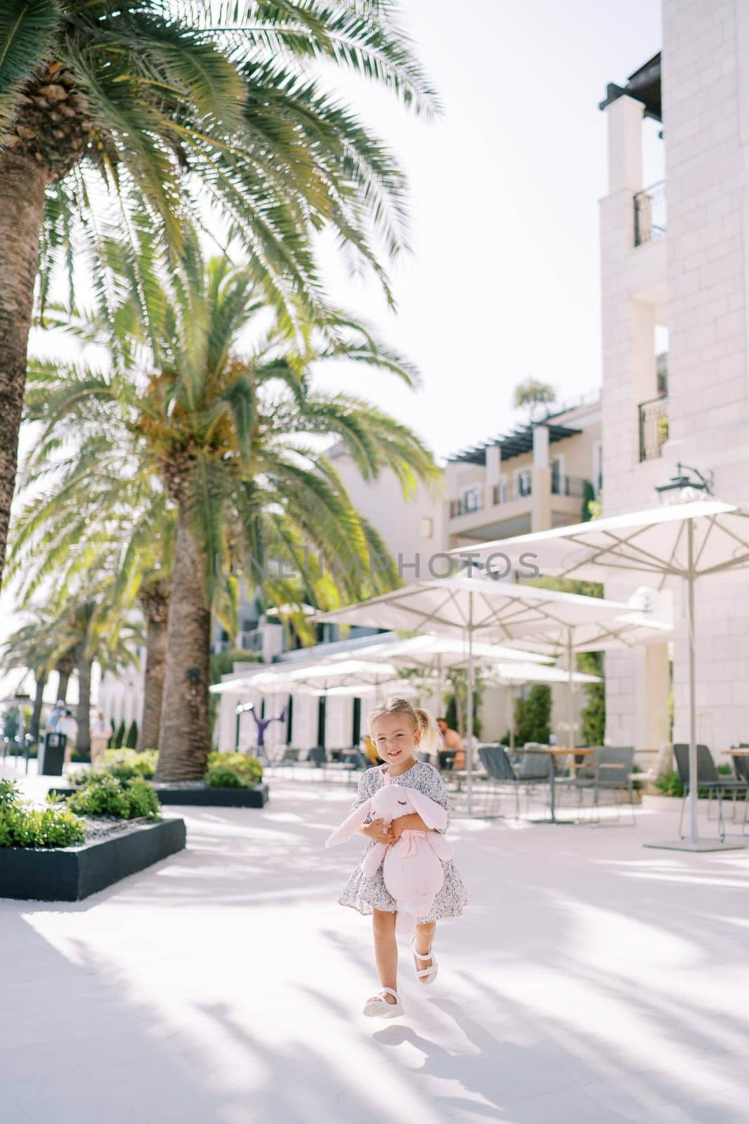 Little girl with a pink toy rabbit runs down the street past green palm trees. High quality photo