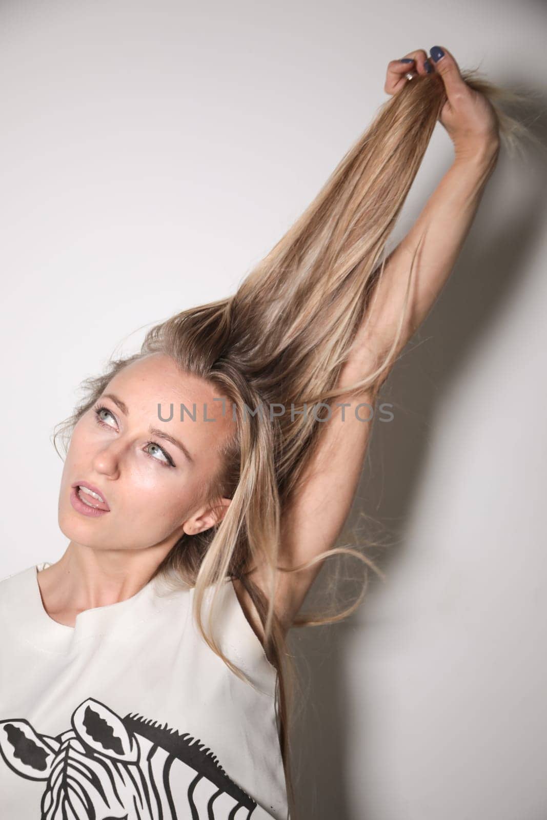 A young model, blonde, dressed in a T-shirt with trimmed sleeves, placed in front of a white wall and illuminated by a strong flash light, stretches her hair with one hand.