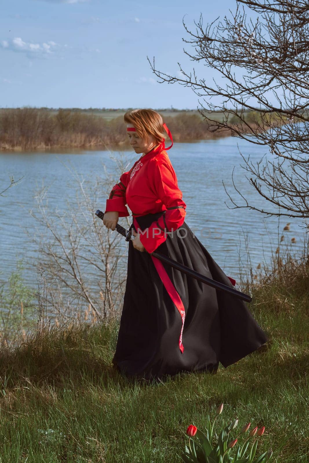 A woman with short red hair in black and red hanfu and a katana on the background of a river in spring
