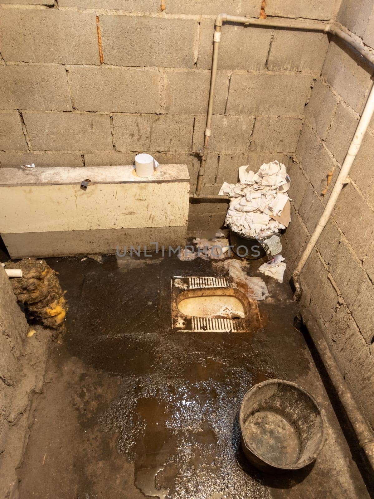 Dirty toilet room with genoa bowl, pile of used toilet paper and a bucket, uncoated concrete flooring and cinder brick walls. Developing country poorness.